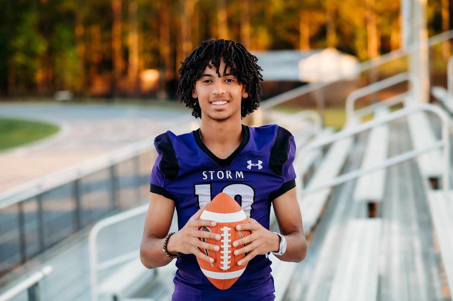 Young football player in purple Storm jersey holding a football, smiling at stadium bleachers during golden hour.