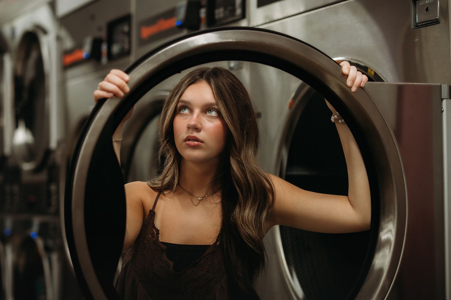 Young woman with long brown hair posing inside a laundromat washing machine door, wearing a dark top.