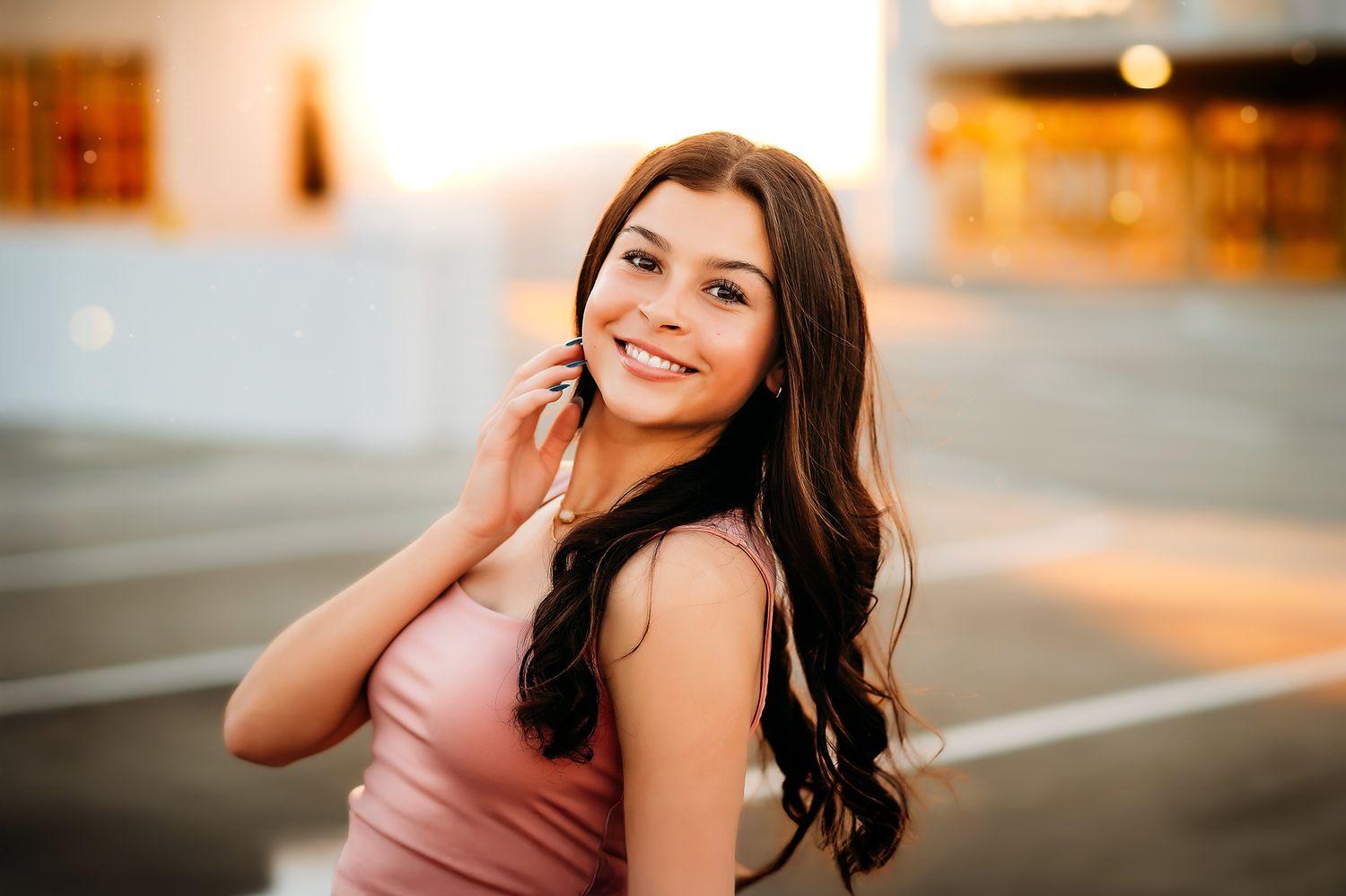 Smiling young woman with long dark wavy hair talking on phone outdoors in warm golden light wearing pink tank top.