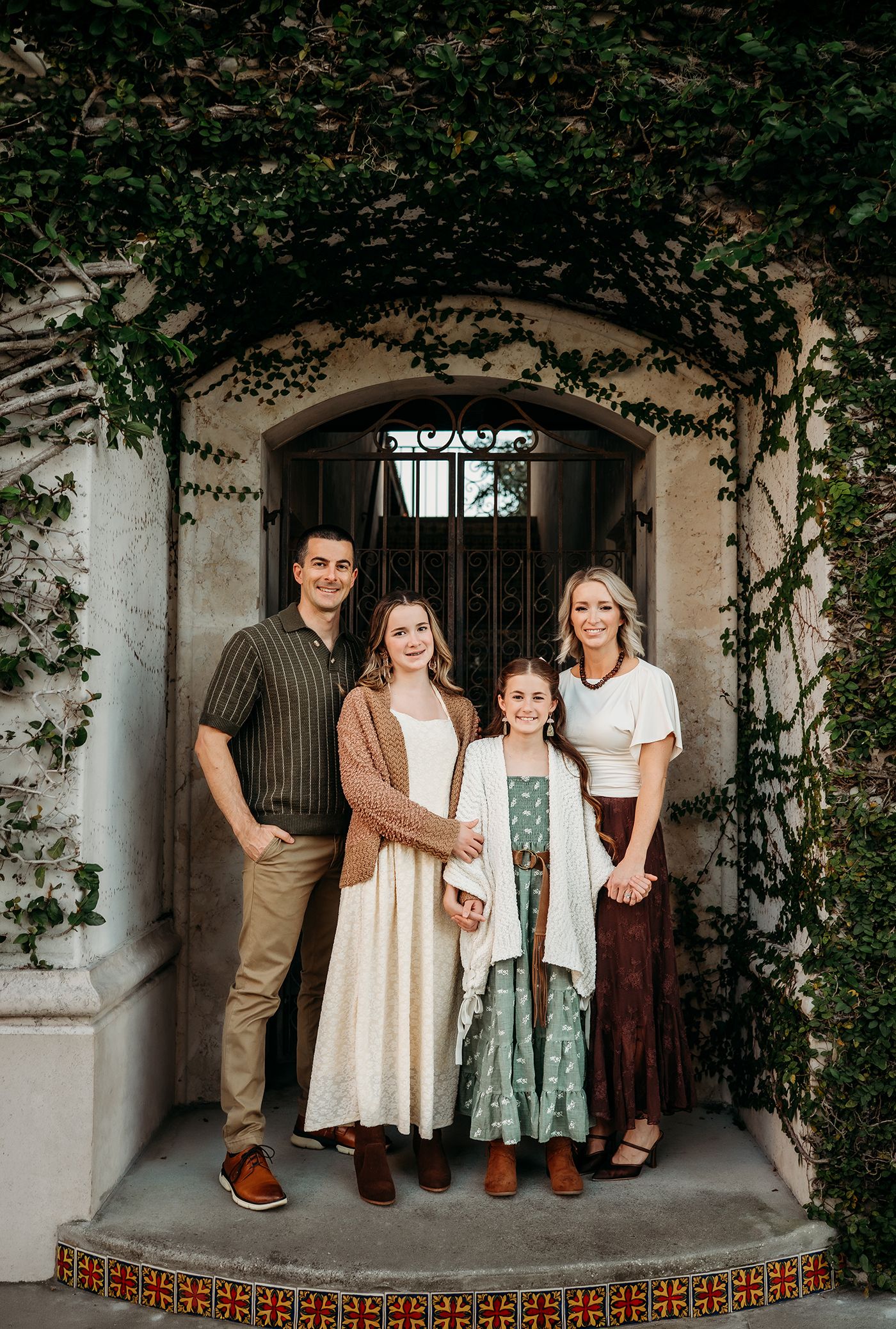 Family of four posing on tiled steps beneath a vine-covered stone archway with an iron gate, in coordinated earth-tone outfits.