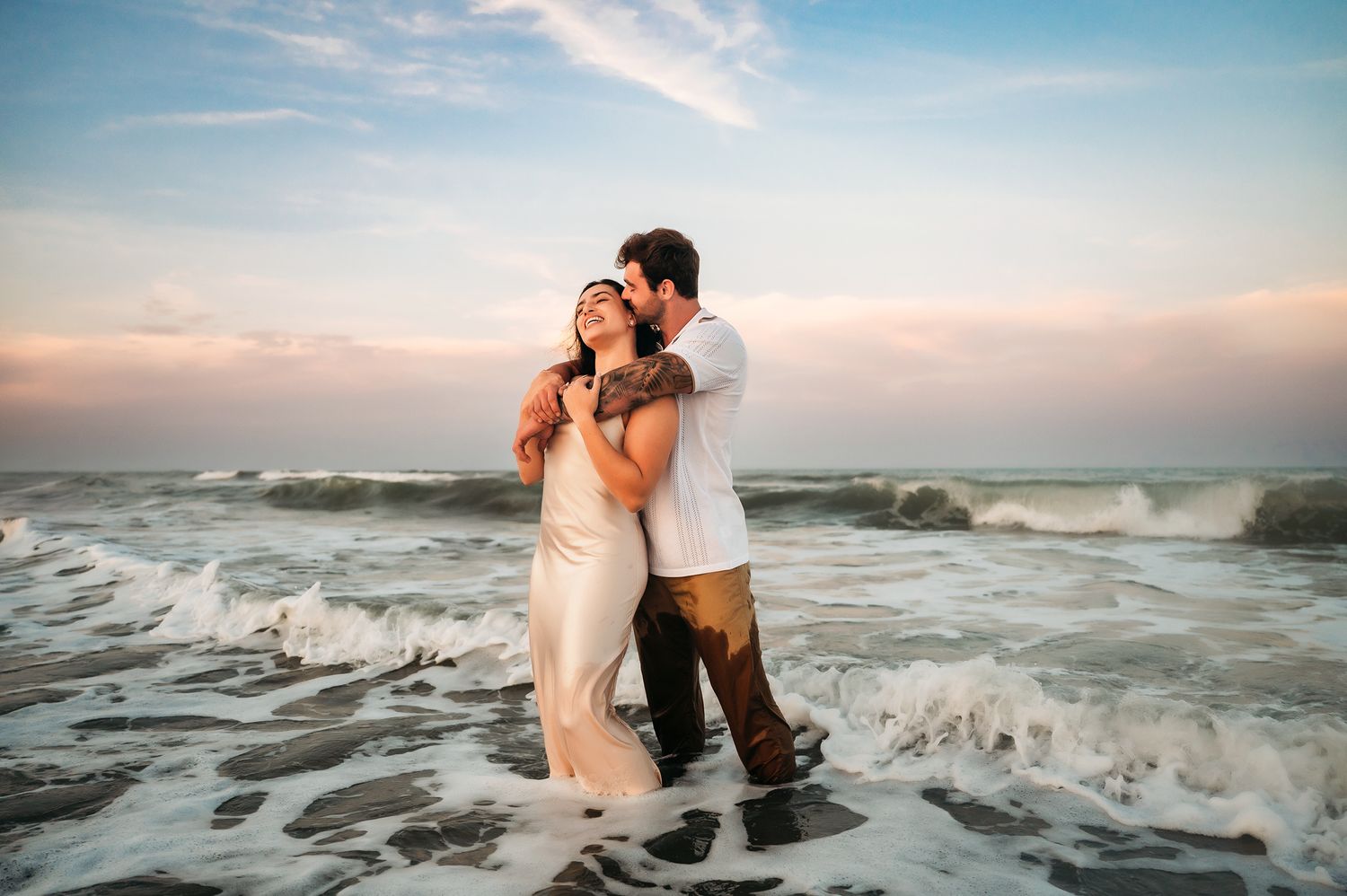 Couple embracing romantically on a beach at sunset with ocean waves crashing around them.