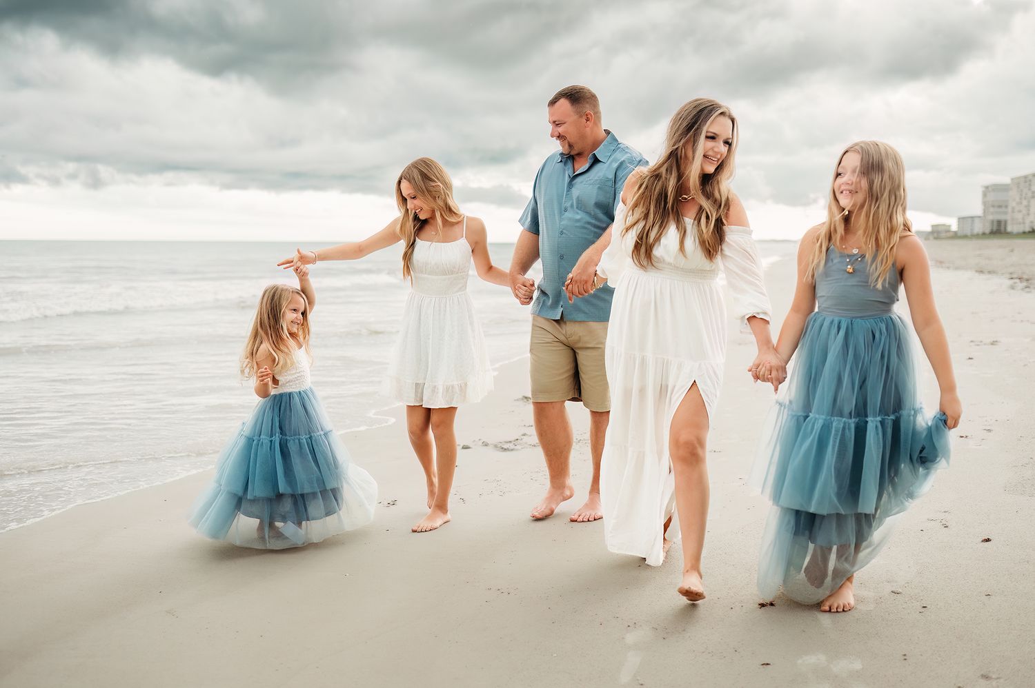 Family of five walking on beach, wearing coordinated blue and white outfits, holding hands near ocean waves.
