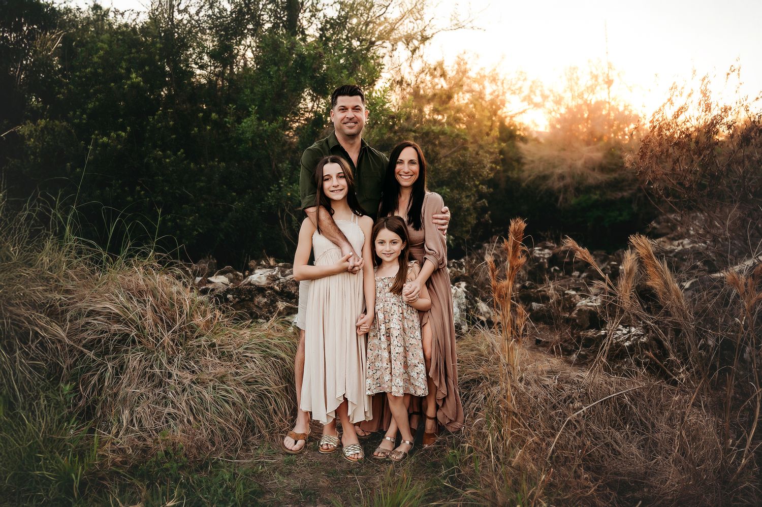 Family of four poses outdoors at golden hour in a field, wearing coordinated boho outfits, smiling warmly together.