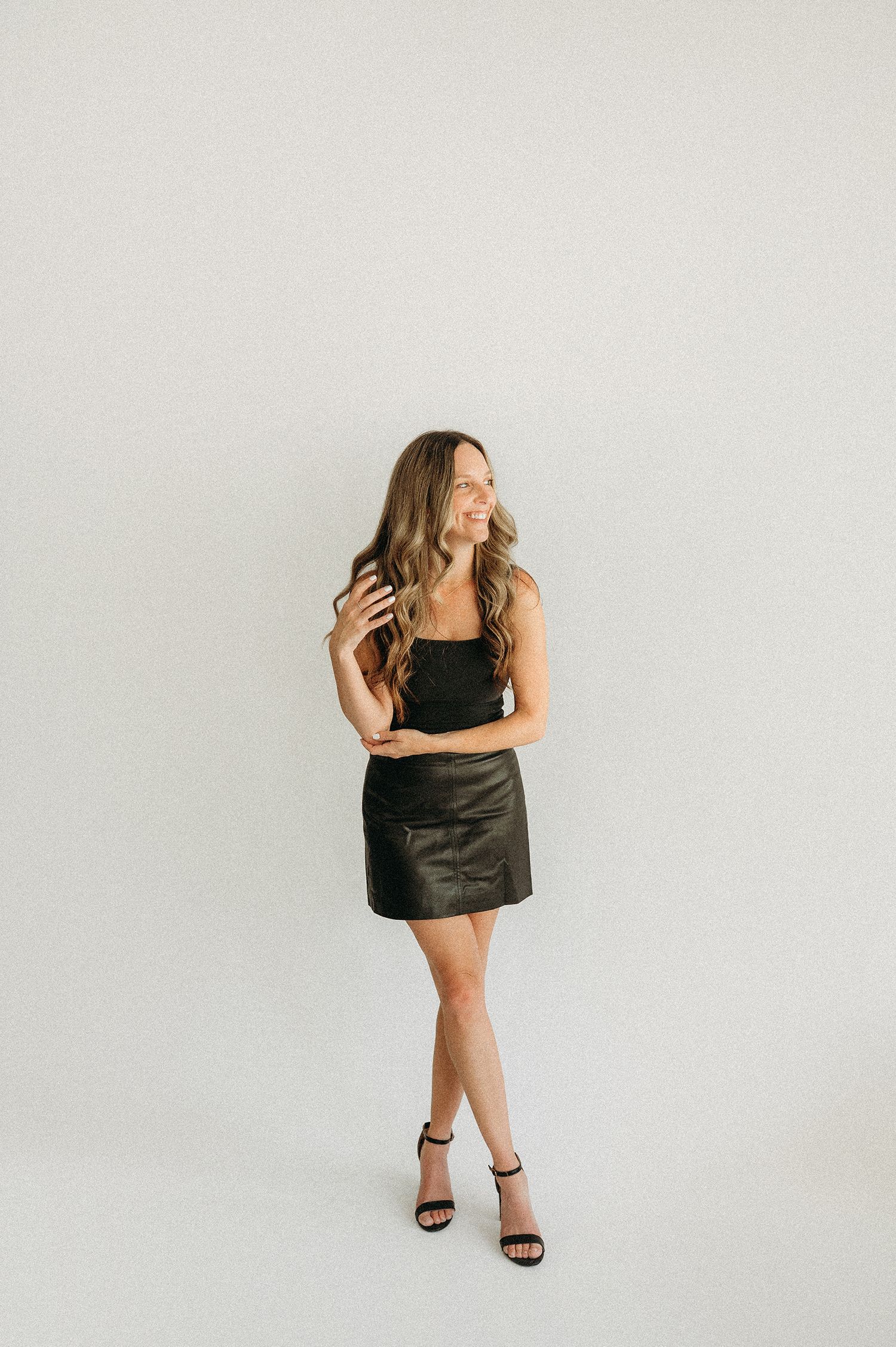 Woman in black crop top and faux leather mini skirt with strappy heels poses against white background.