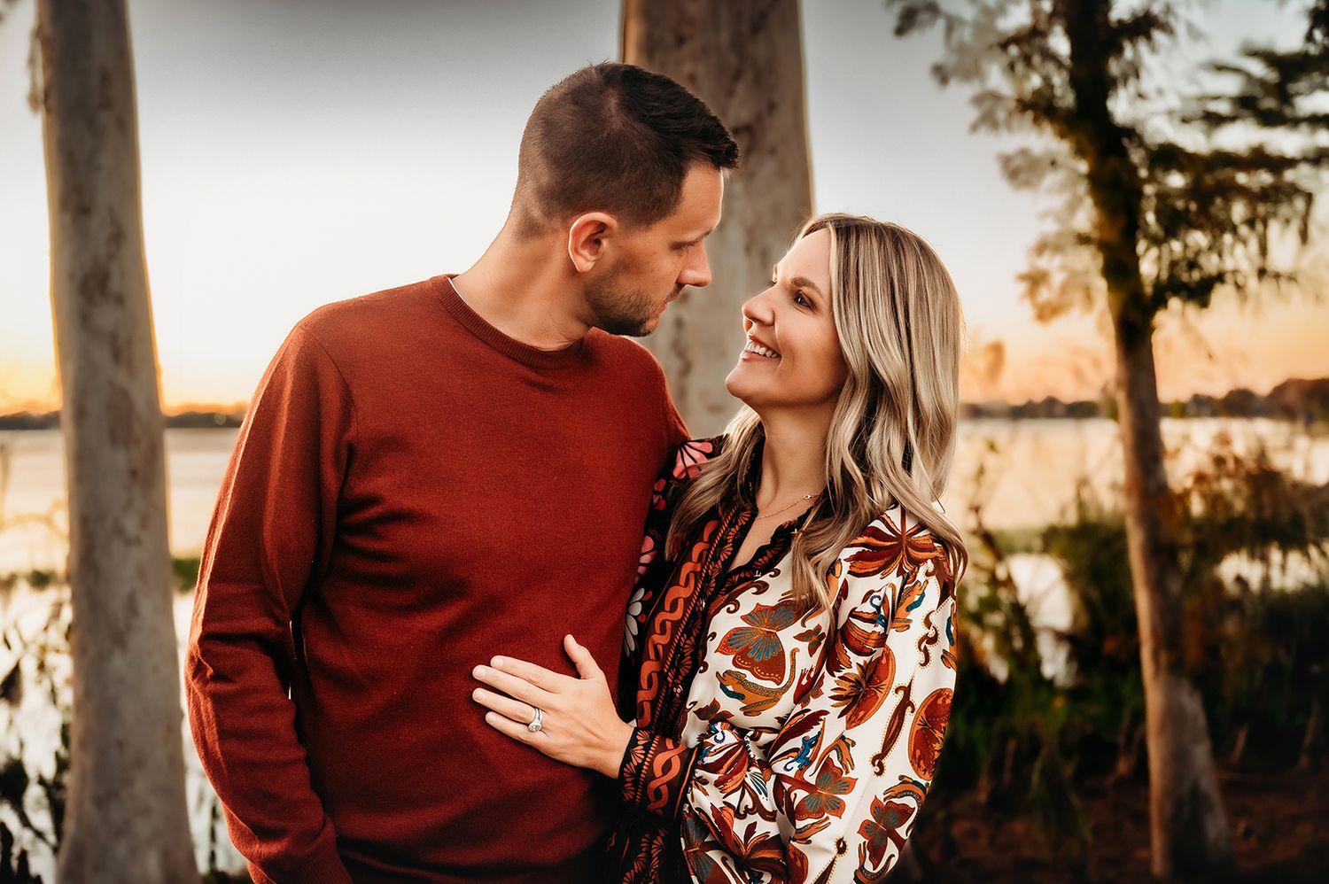 Couple sharing a romantic moment outdoors near a lake, man in red sweater, woman in floral blouse, golden hour light.