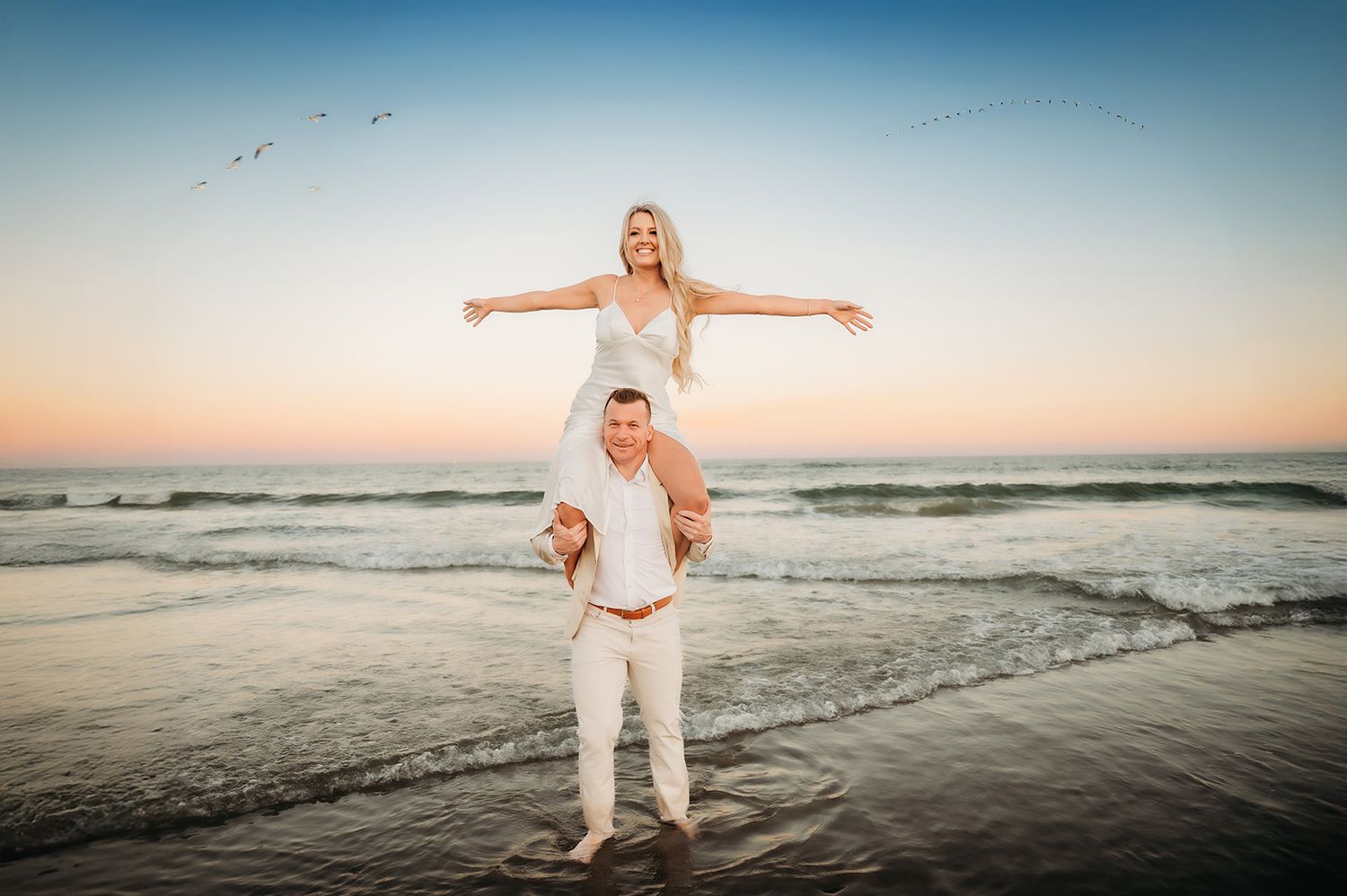 Couple in white outfits on beach at sunset, woman sitting on mans shoulders with arms outstretched by ocean waves.