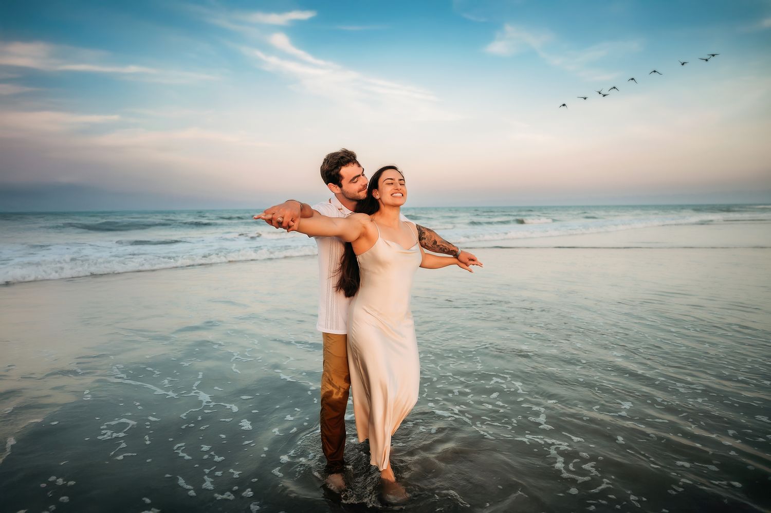 Couple embracing on a beach at sunset, woman in white dress with arms outstretched, waves and pastel sky behind them.
