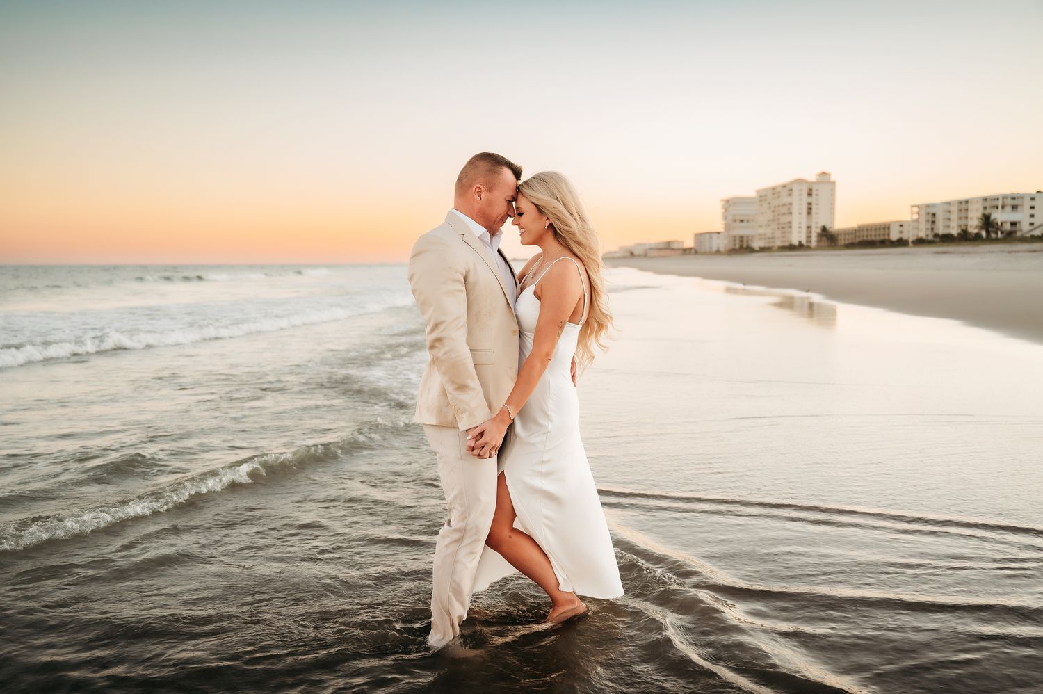 Couple in white attire sharing a romantic kiss on a sandy beach at sunset with ocean waves around their feet.