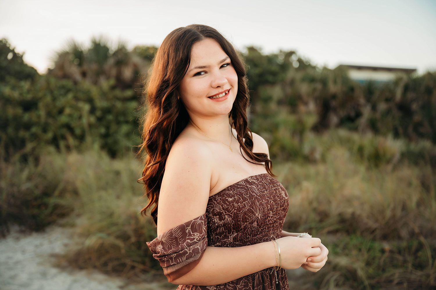 Young woman in brown off-shoulder dress smiling outdoors near beach dunes with lush greenery background.