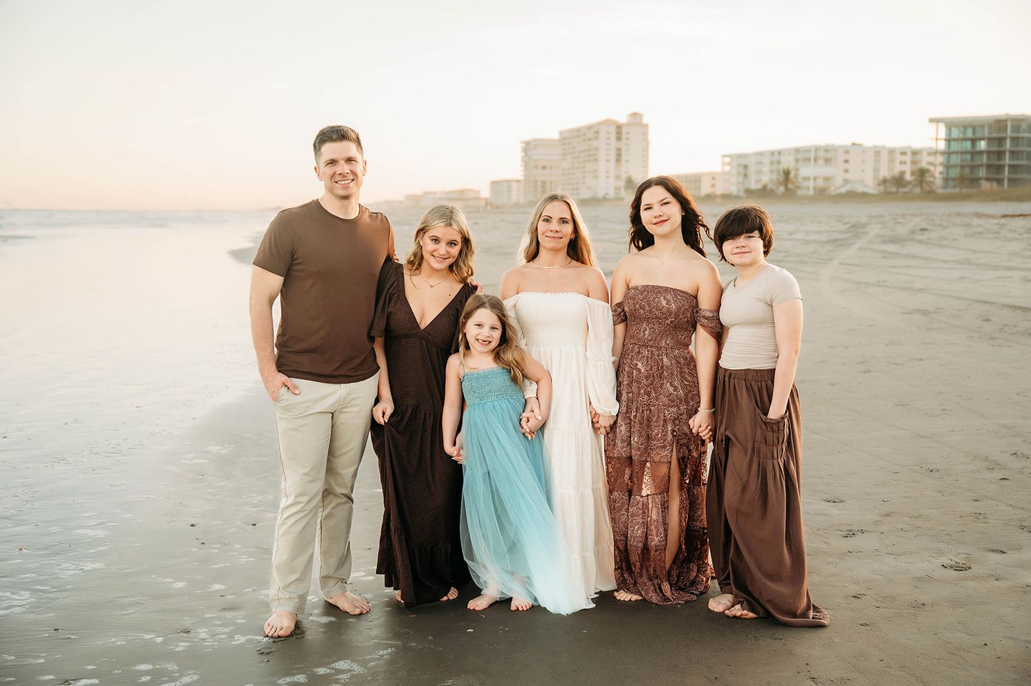 Family of six posing together on a beach at sunset, wearing elegant dresses and casual attire, with ocean waves and buildings behind.