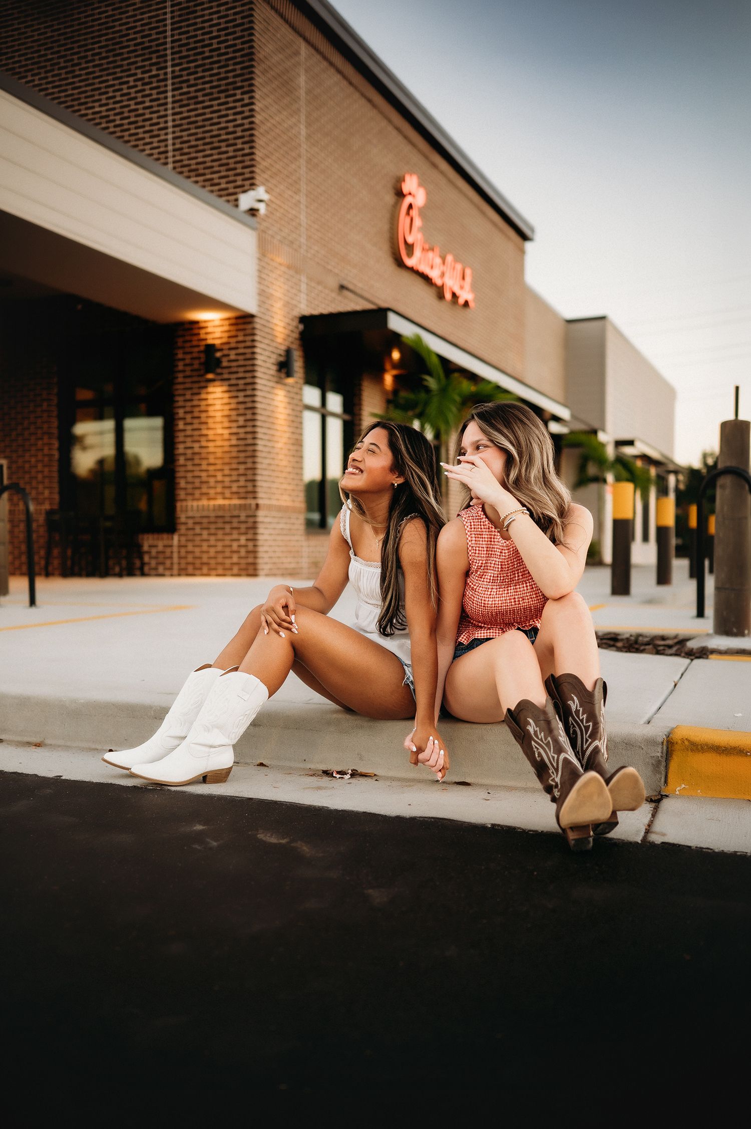Two women in cowboy boots sit laughing on a curb outside a brick Chick-fil-A restaurant at golden hour.