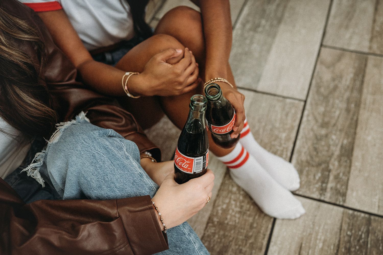 Two people sitting on floor clinking Coca-Cola glass bottles together, wearing casual outfits with jeans and white socks.