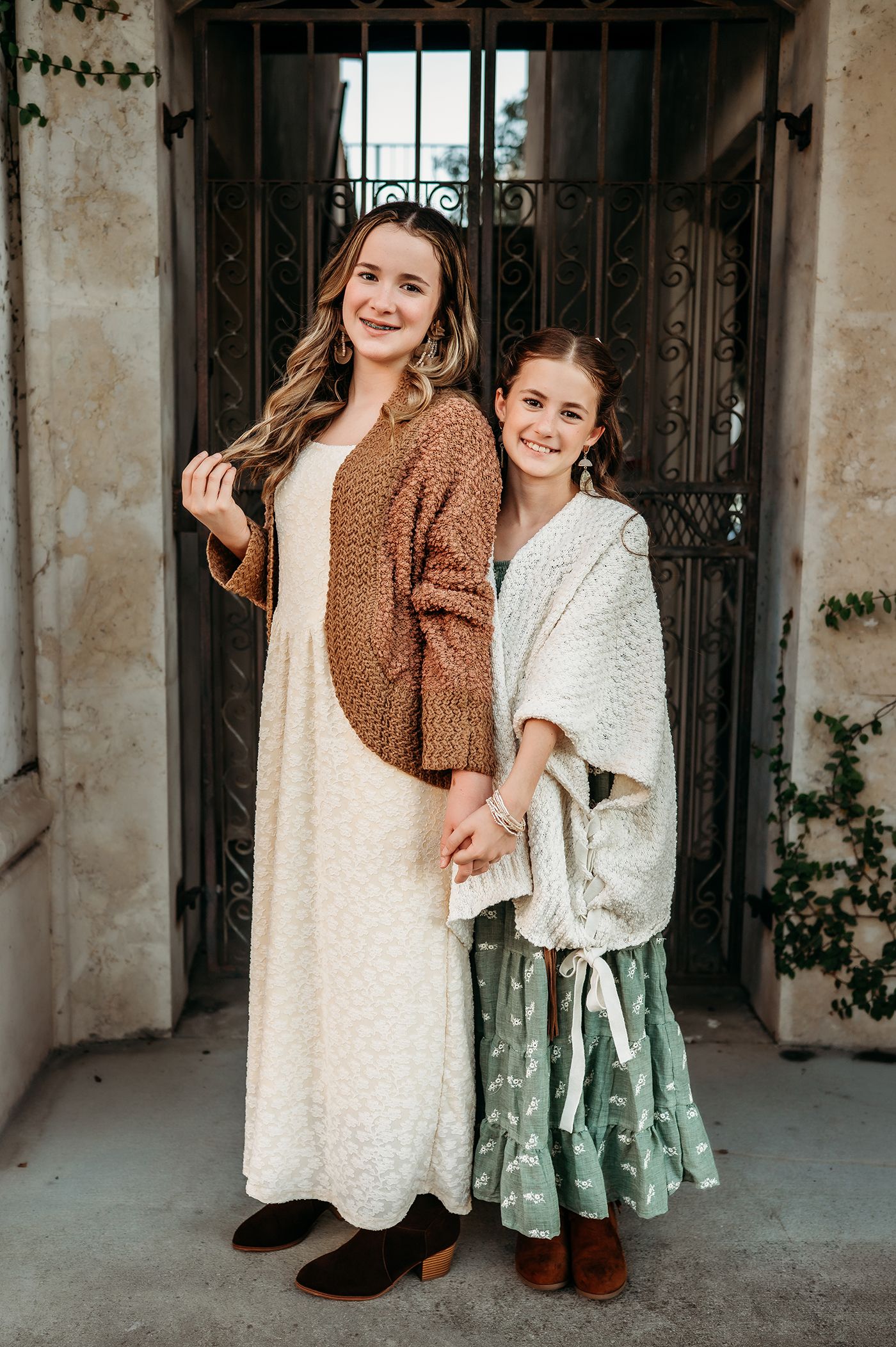 Two smiling girls in cream and sage boho dresses with knit cardigans pose hand in hand before an ornate wrought iron gate.