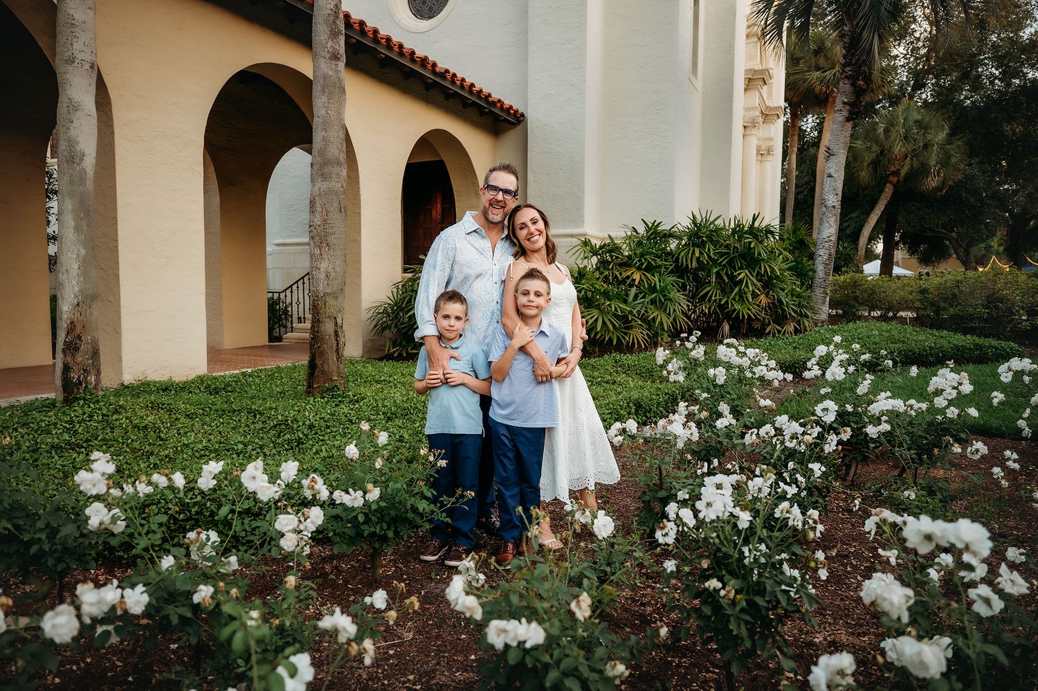 Family of four posing in garden with white flowers outside arched building with palm trees.
