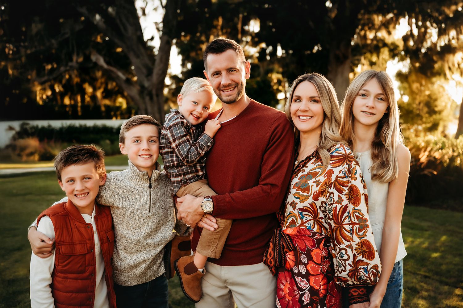Happy family of six posing outdoors at golden hour with lush green trees in the background.