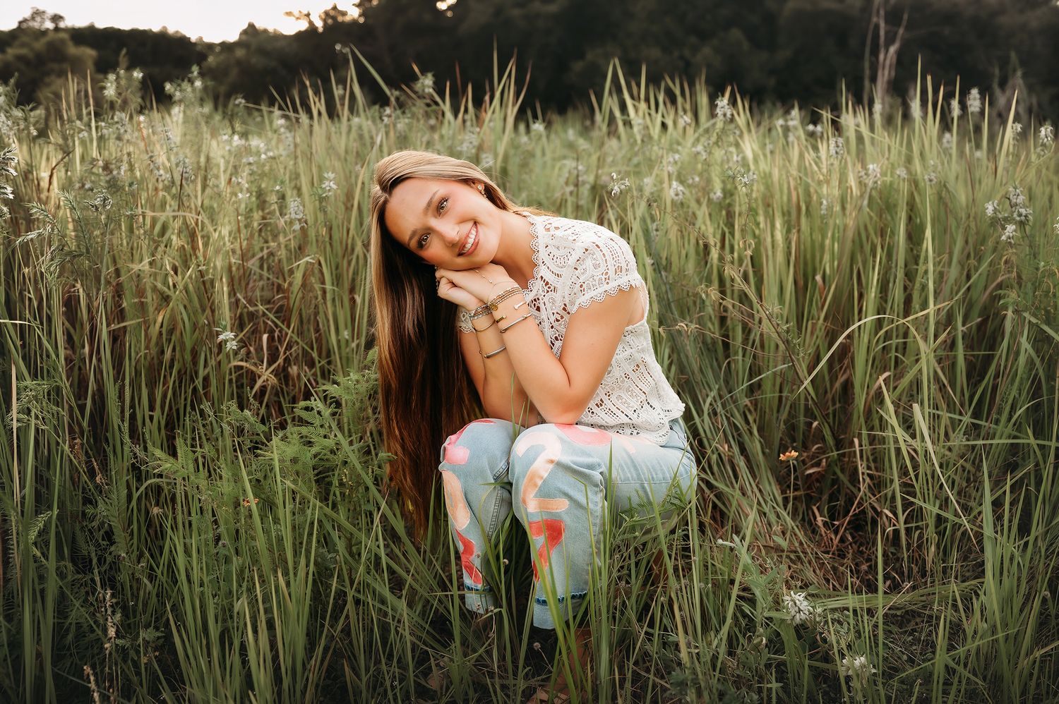 Young woman with long brown hair sitting in tall green grass field, wearing floral pants and white top, smiling softly.