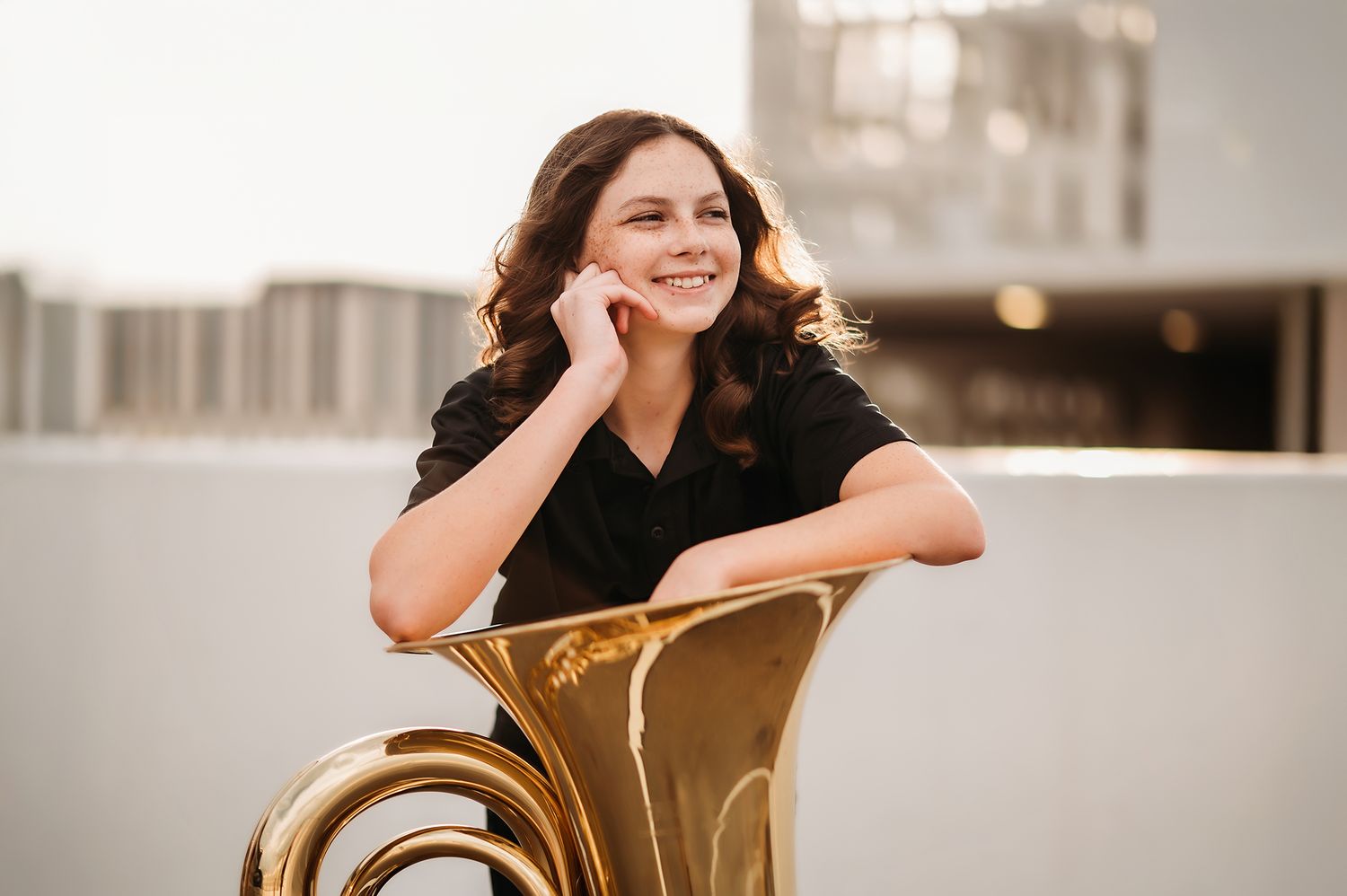 Young woman smiling and leaning on a gold tuba during an outdoor senior portrait session.