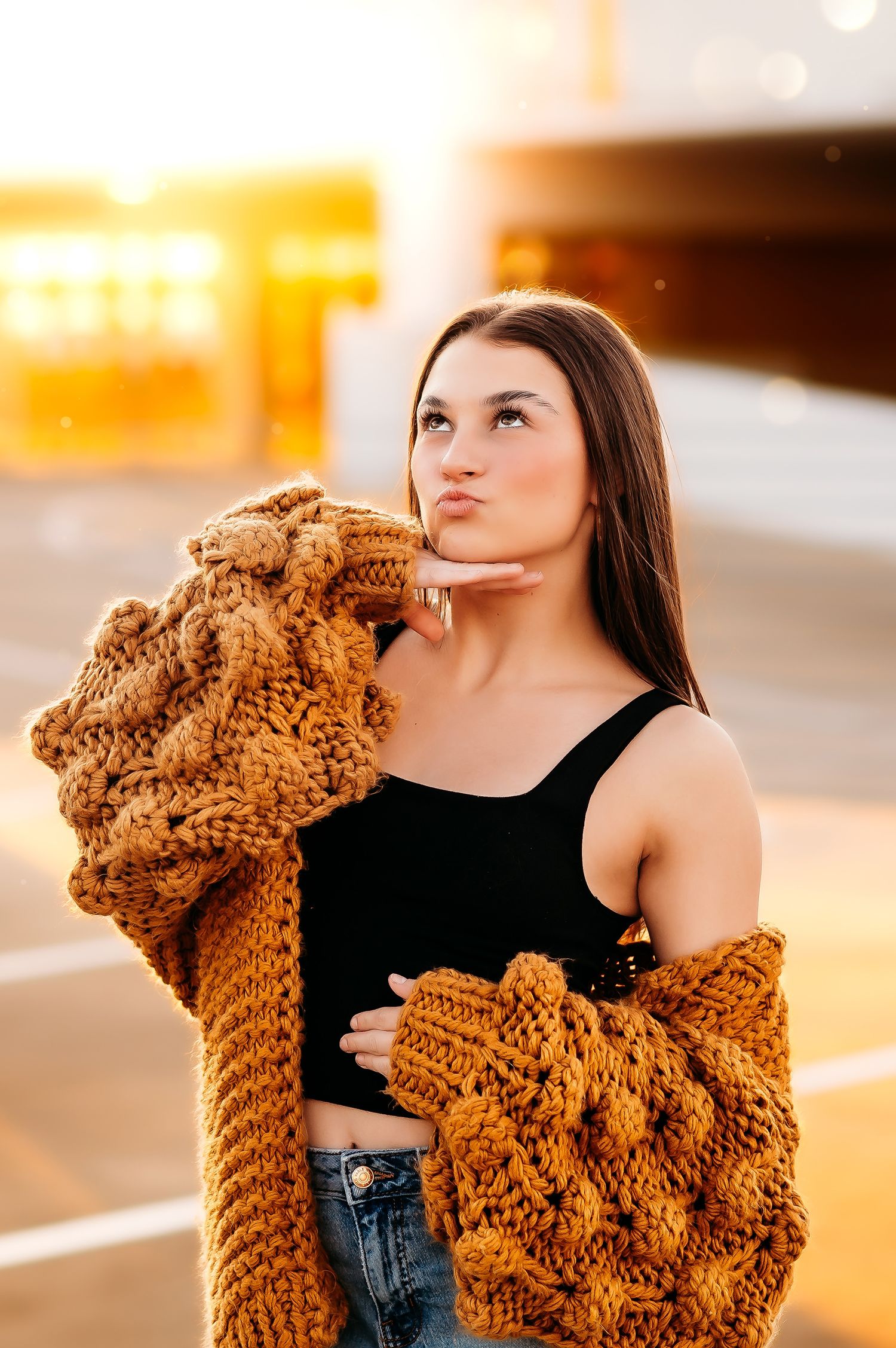 Young woman in chunky knit mustard cardigan and black crop top posing outdoors in golden sunset light.
