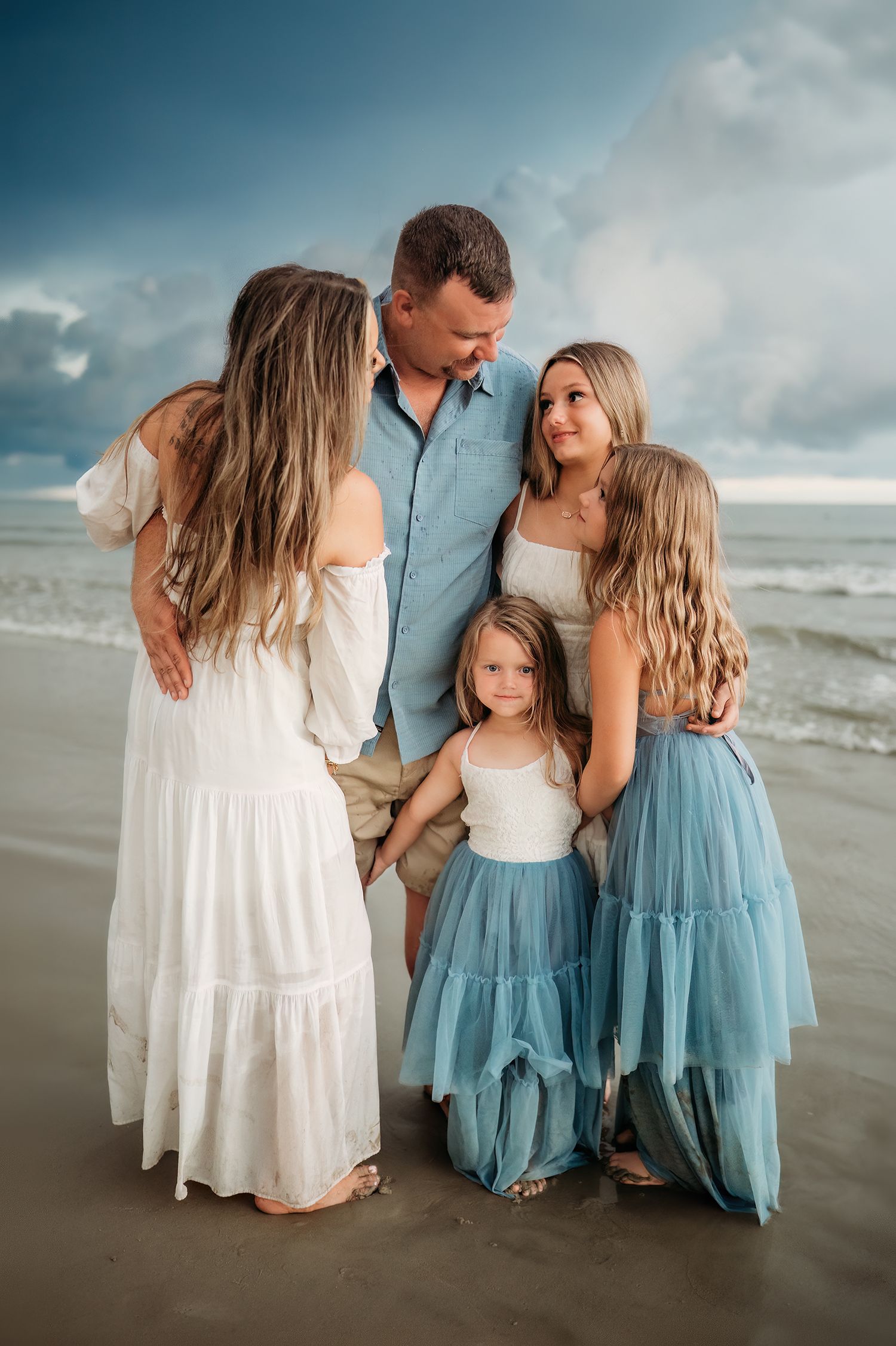 Family of five posing together on a beach, wearing white and blue outfits, with ocean waves and cloudy sky behind them.