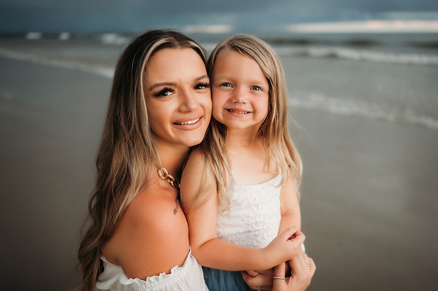 A smiling young woman hugs a little blonde girl on a beach, both wearing white, with ocean waves in the background.