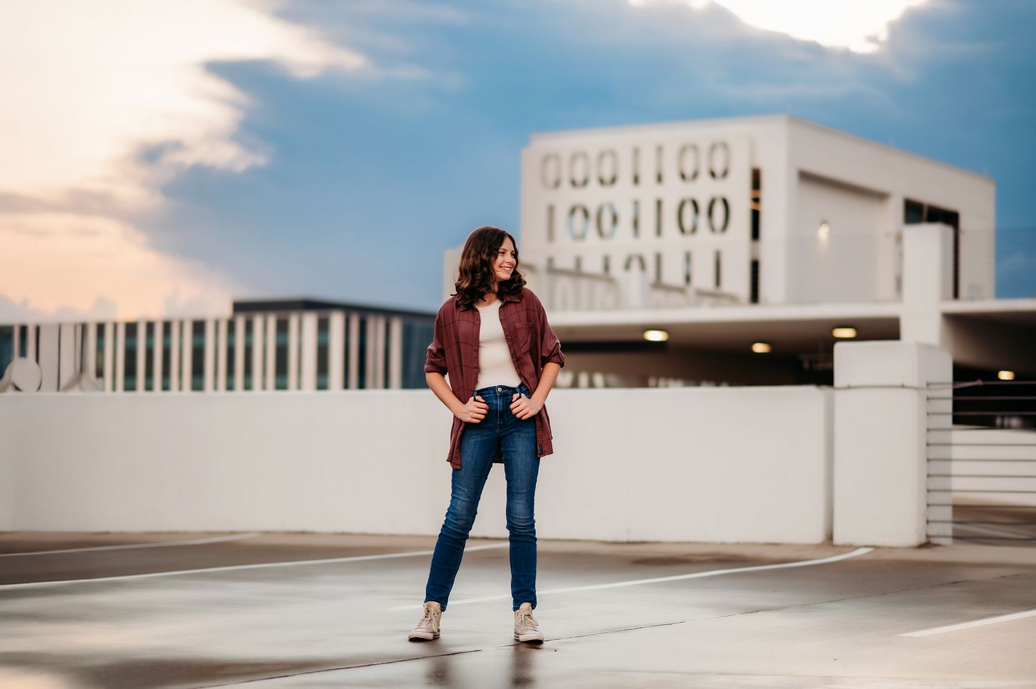 Young woman posing confidently on a rooftop parking garage at sunset in jeans and a red jacket.