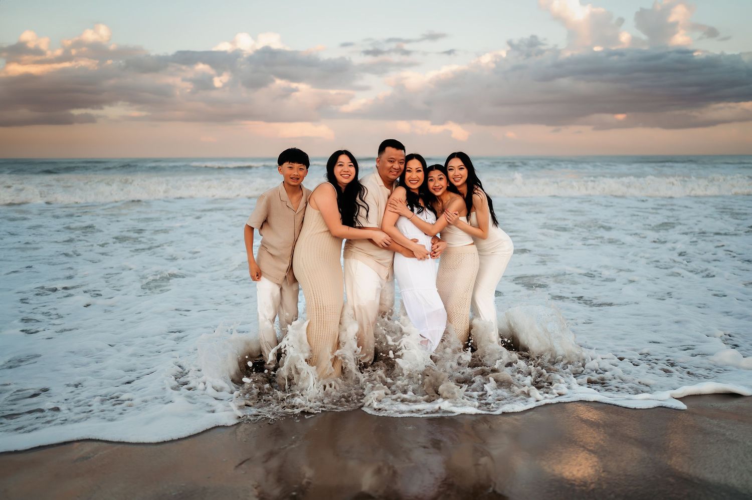 A joyful extended family group embraces in ocean waves at sunset on a tropical beach.