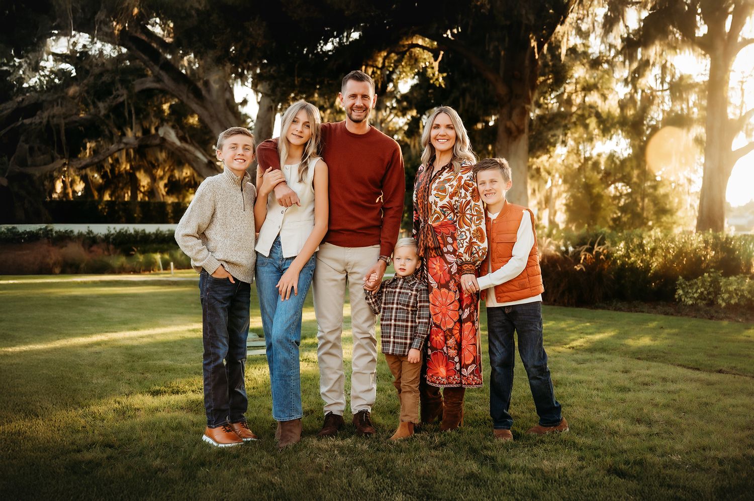 Family of six posing outdoors at golden hour beneath large oak trees on a green lawn.