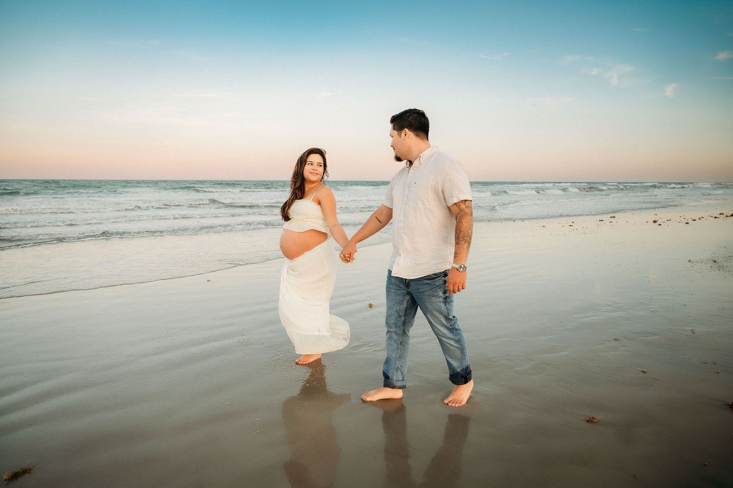 Pregnant woman in white dress holds hands with man walking on beach at sunset.
