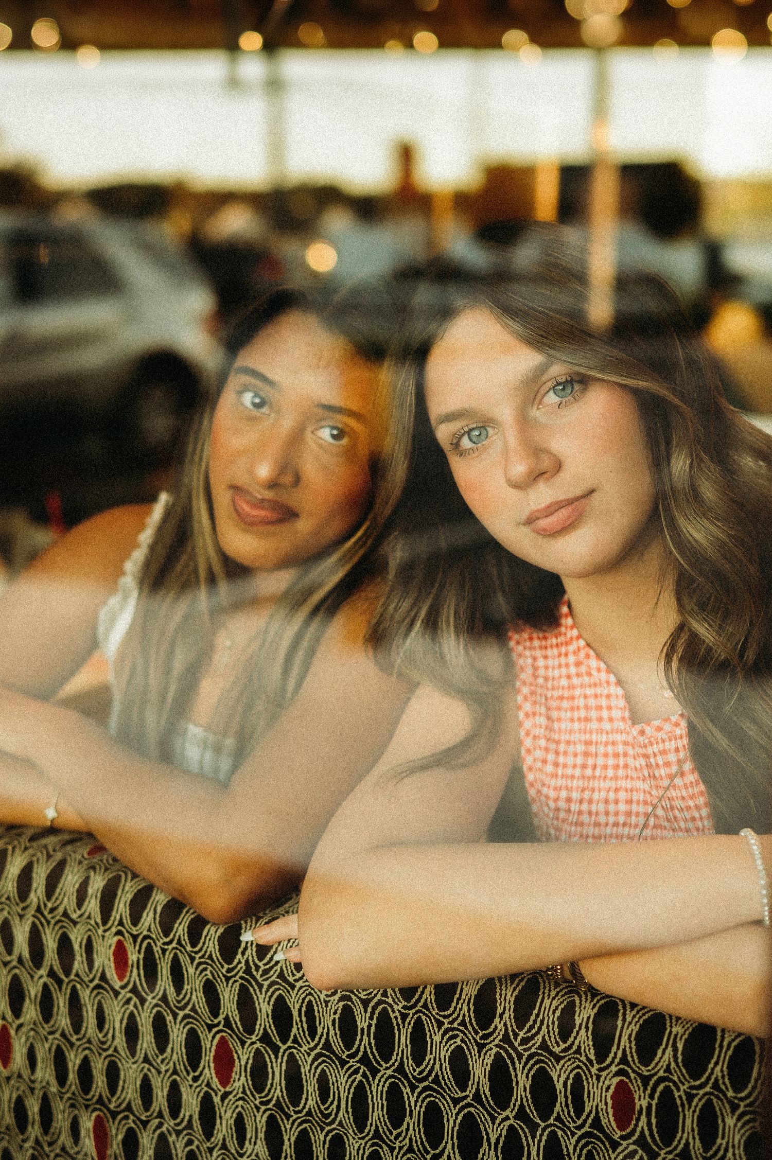 Two young women with long hair lean on a patterned bench, gazing at the camera in a warm, softly lit window reflection.