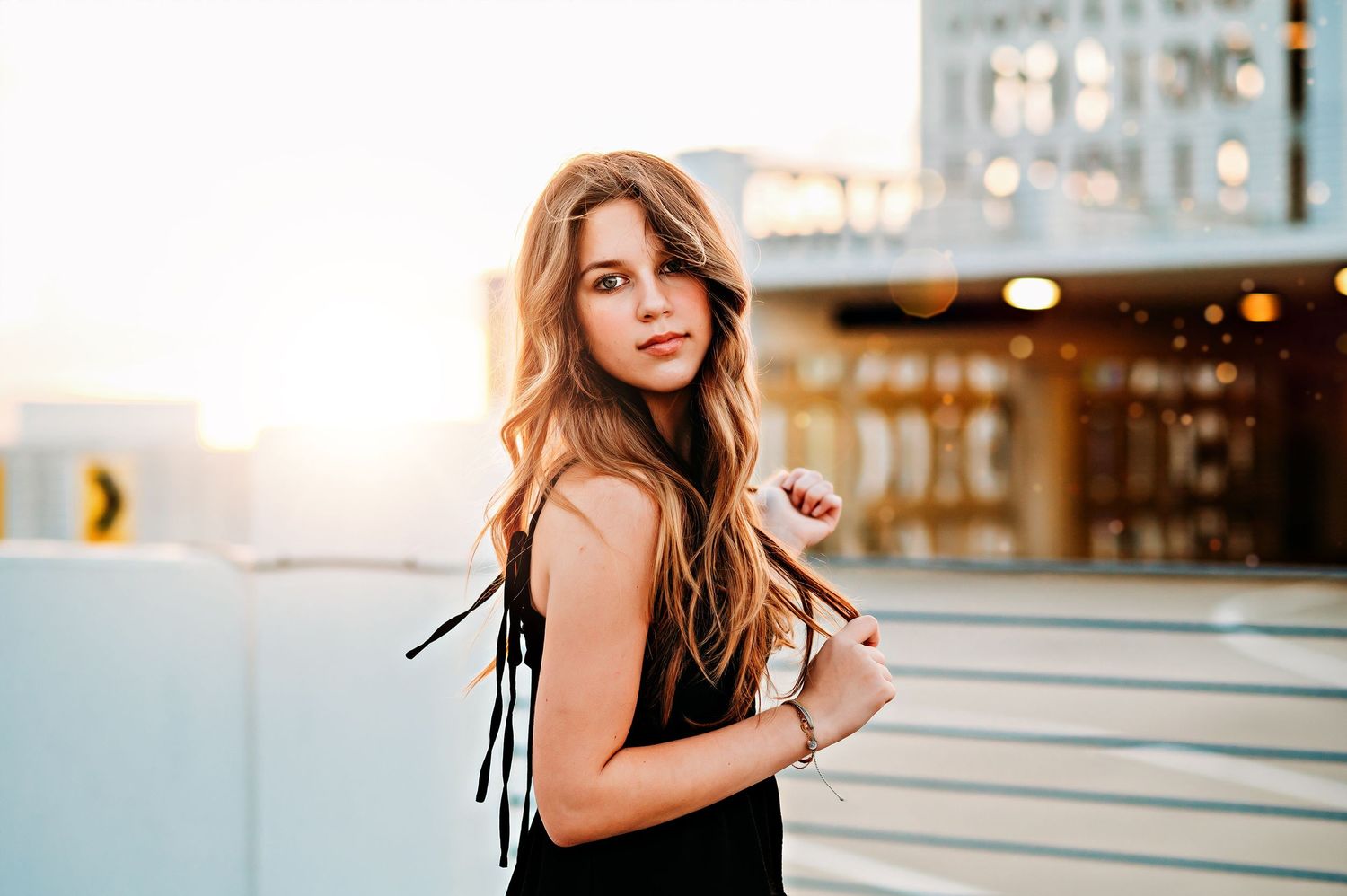 Teen girl with long wavy hair poses in black dress on rooftop parking garage at golden sunset.