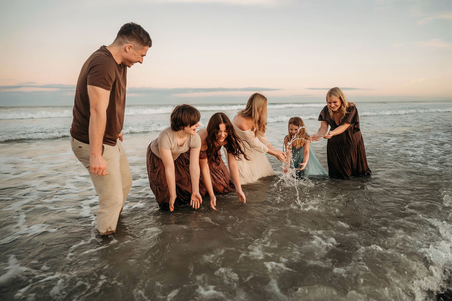 Family of six playing in ocean waves at beach during sunset, wearing earth tones, children splashing water.