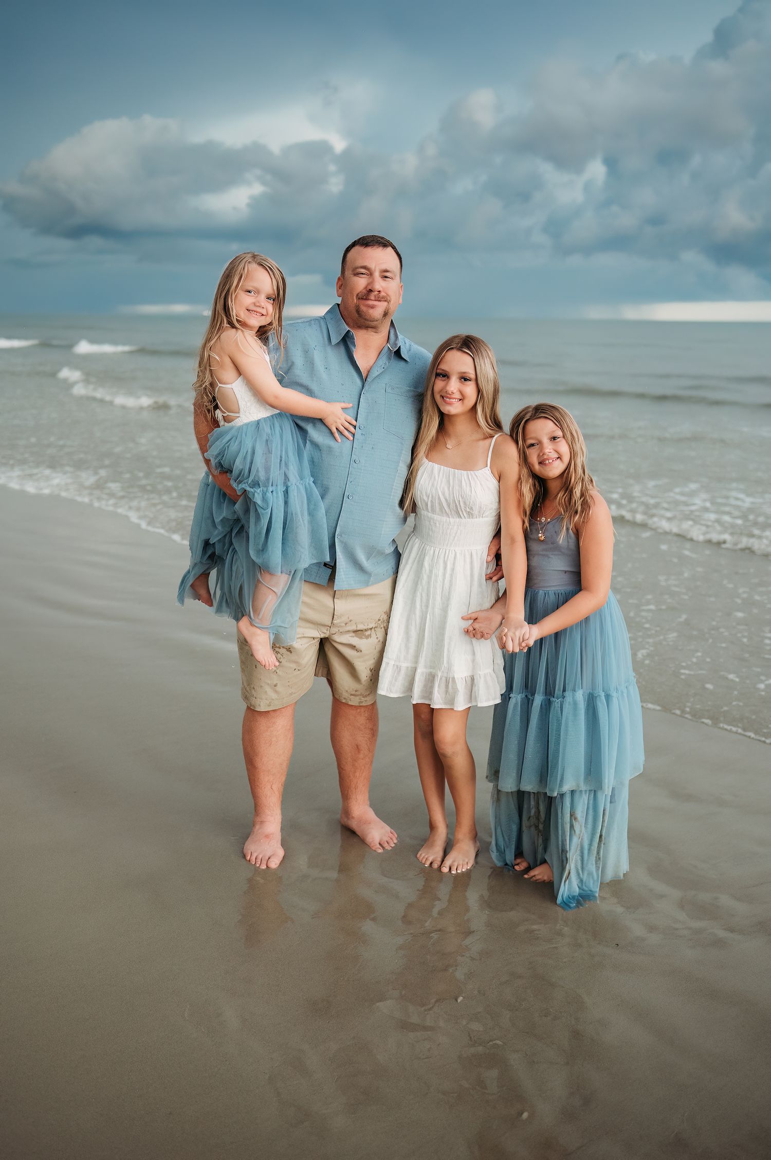 Family of four posing barefoot on a beach, wearing coordinating blue and white outfits, with ocean waves and cloudy sky behind them.