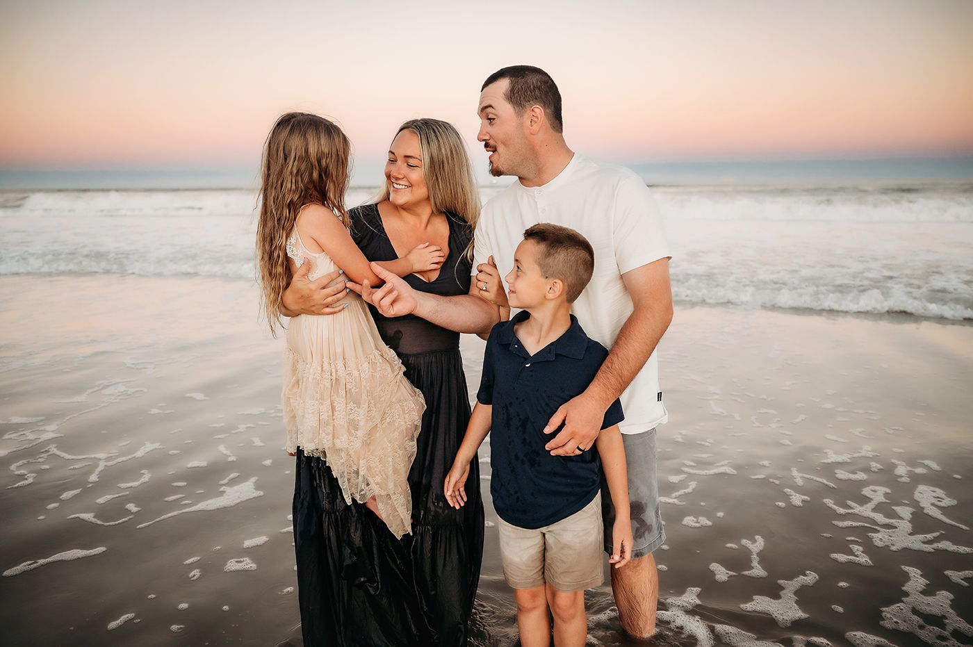 Family of four laughing together on a beach at sunset, waves washing around their feet.