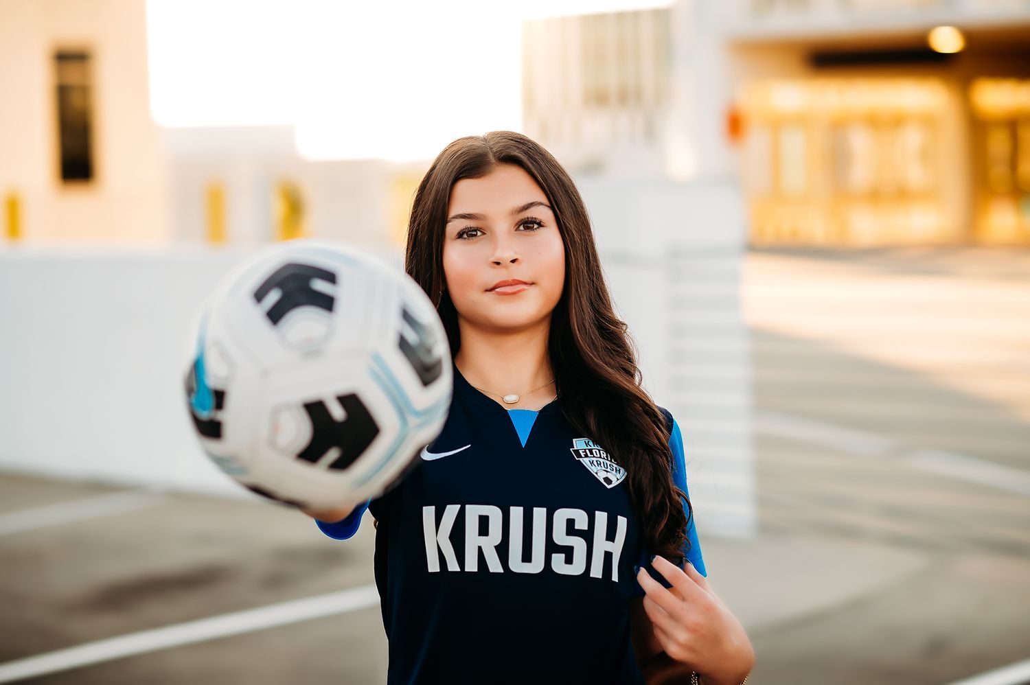 Young female soccer player in Krush jersey balancing a soccer ball outdoors during golden hour.