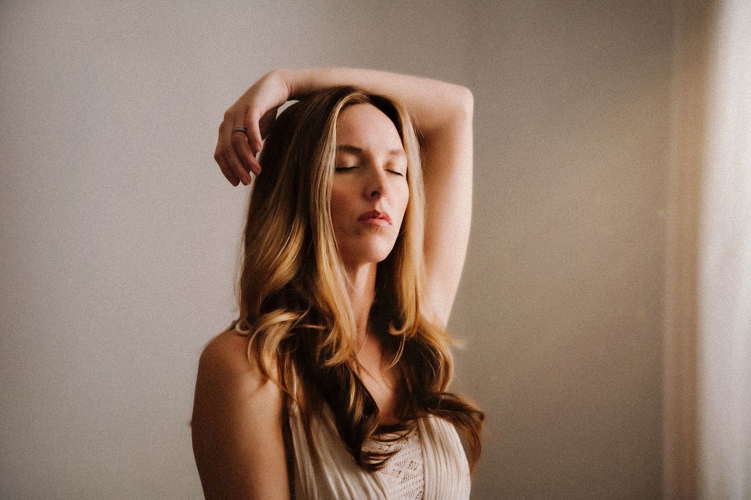 Blonde woman with eyes closed, arms raised gracefully above head, wearing cream dress against neutral background.