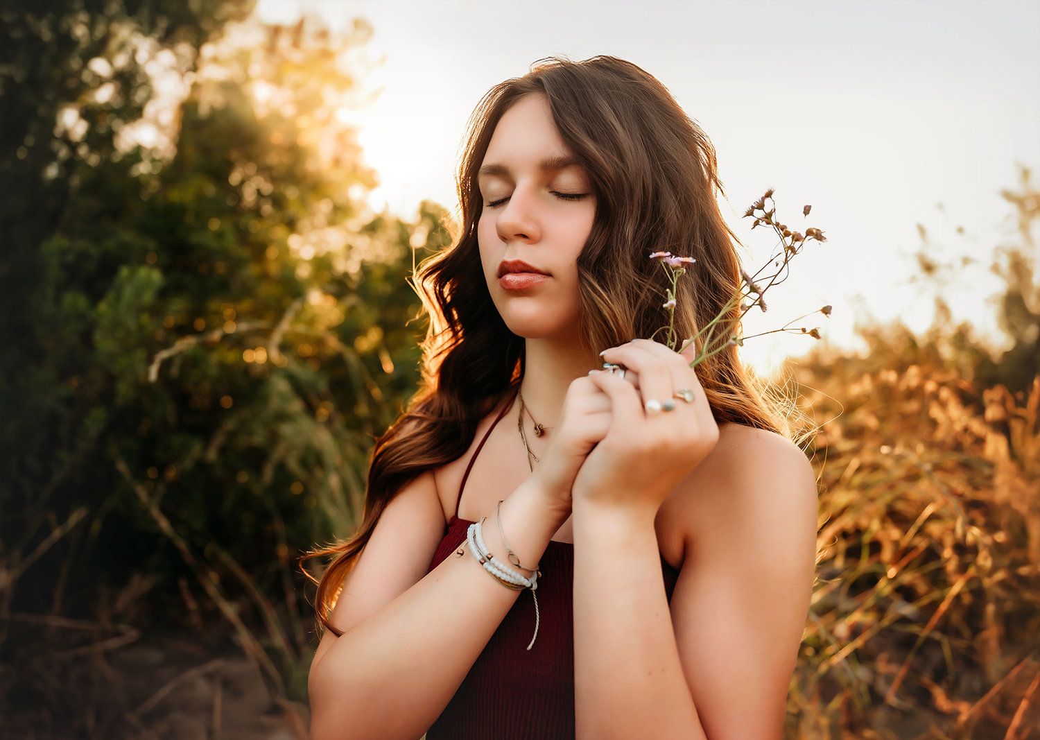 Young woman with closed eyes holding wildflowers in golden sunset light outdoors.