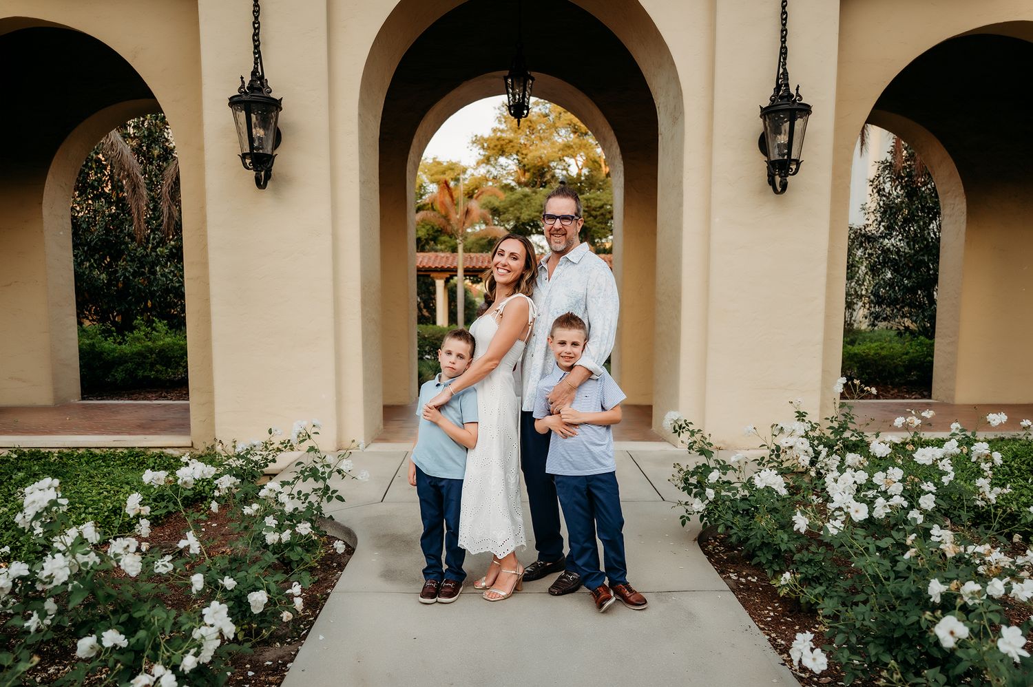 Family of four posing under arched walkway with white roses, parents and two young boys smiling outdoors.