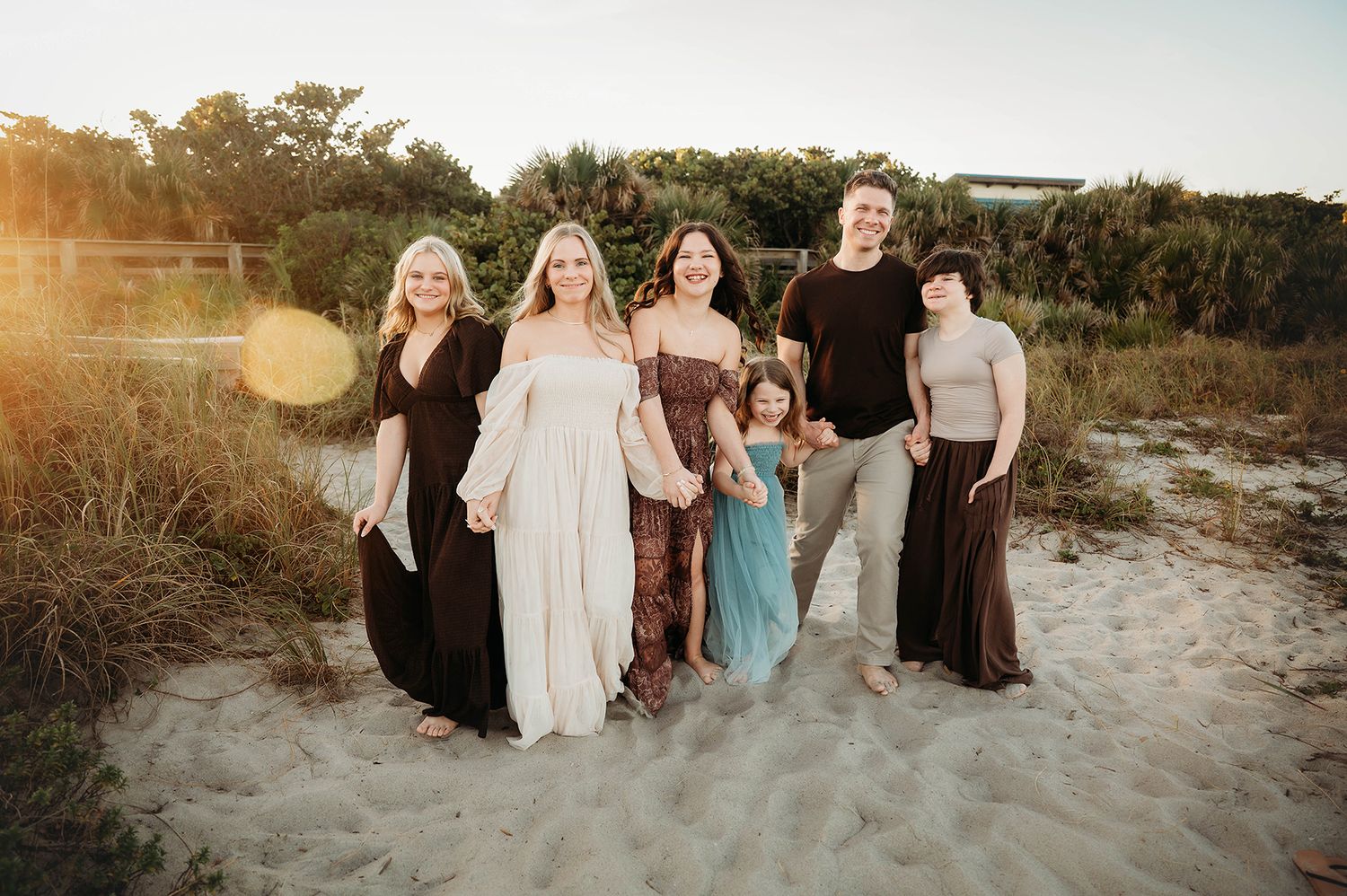 A joyful family of six poses together on a sandy beach at golden hour with lush greenery in the background.