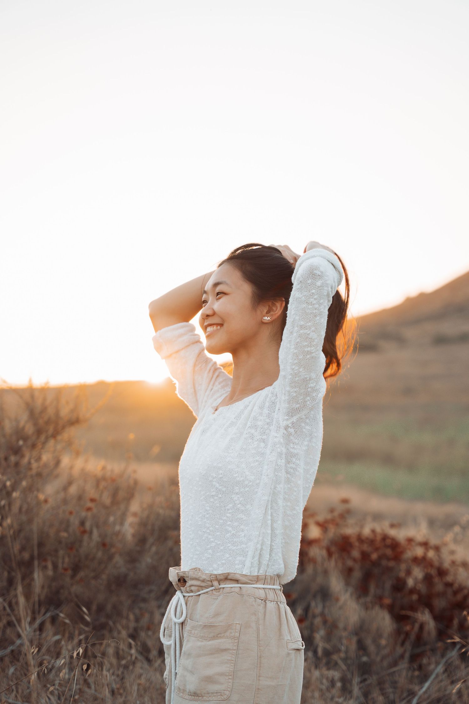 Happy young woman with hands in hair, bathed in warm sunset light in an open field.