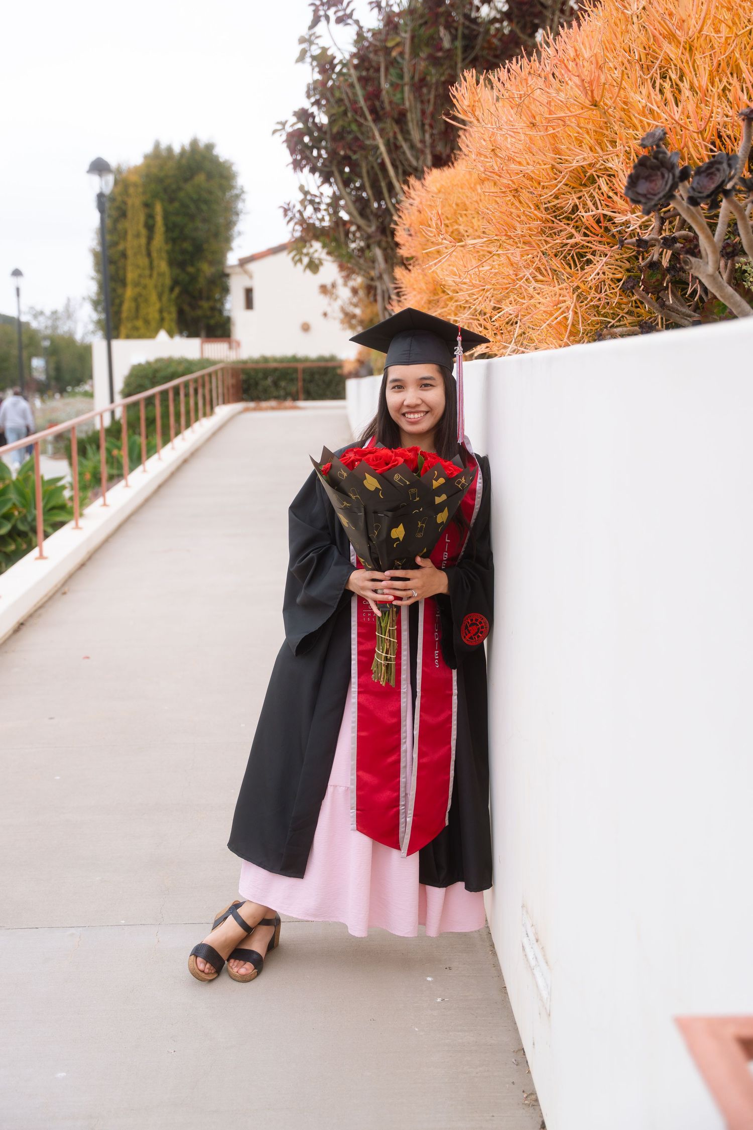Graduate in black cap and gown holding red roses bouquet leaning against white wall outdoors.