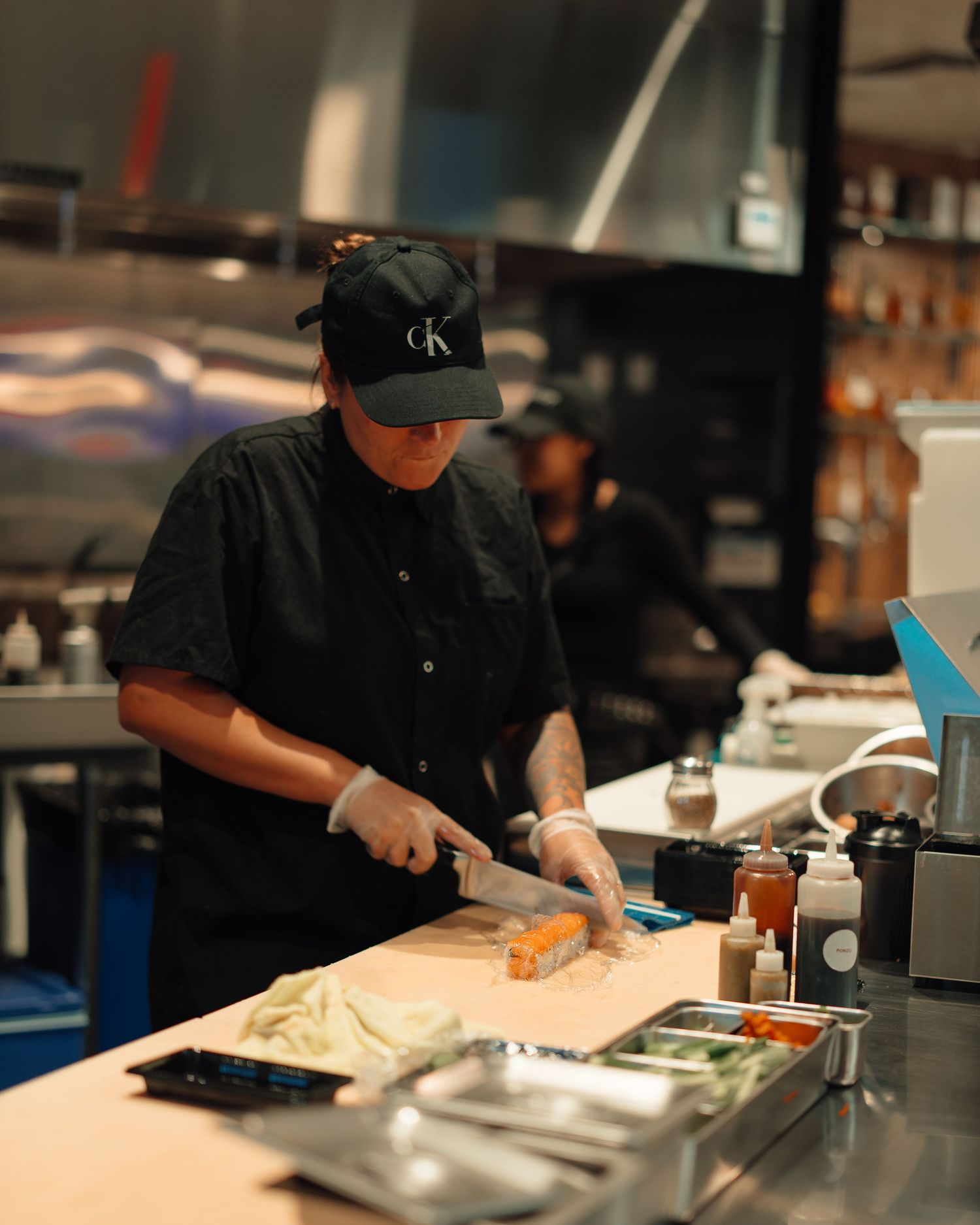 Chef in black uniform and cap cutting food on a wooden board in a professional kitchen.