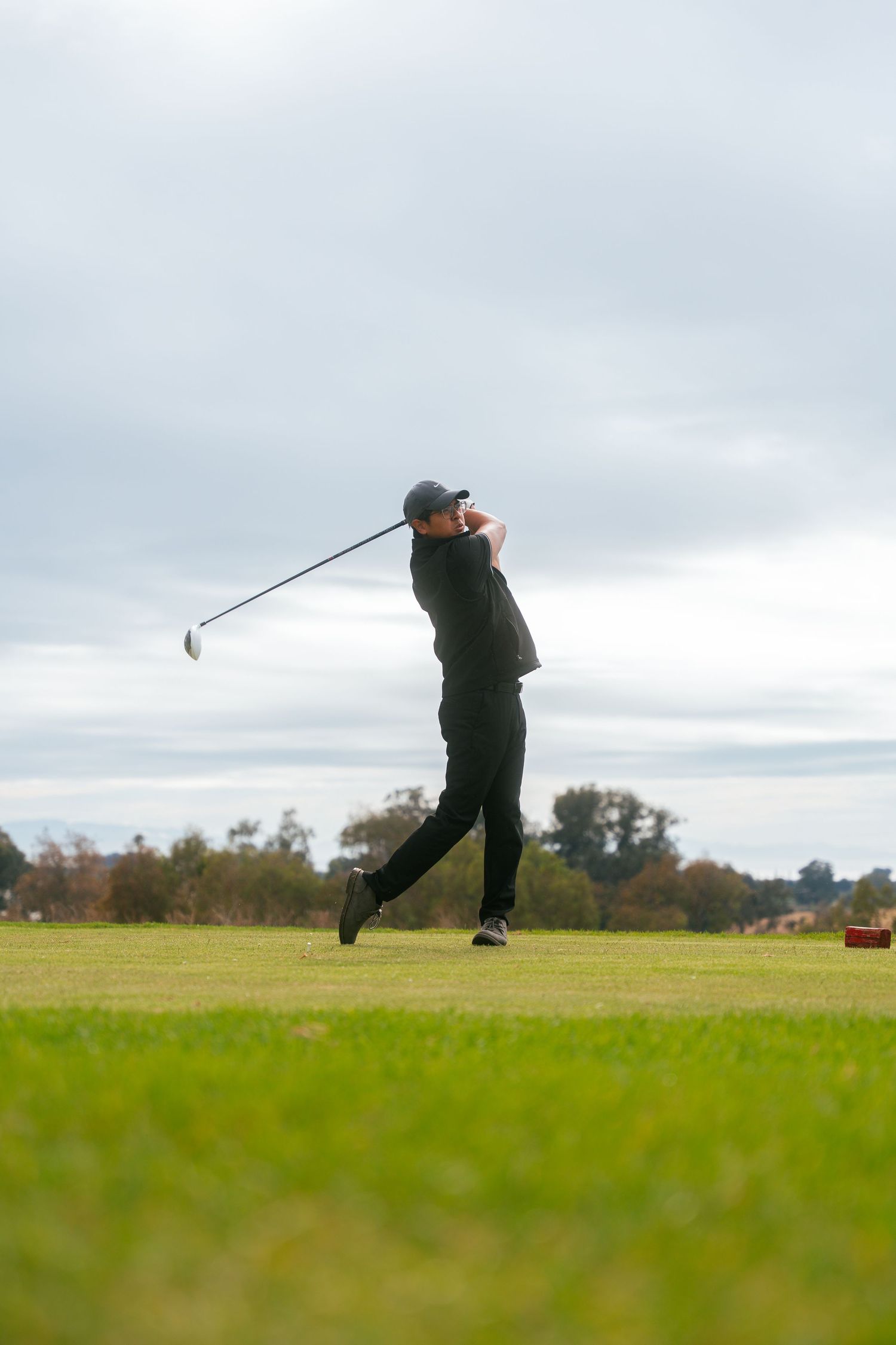 Golfer in black attire completing a full swing on a lush green golf course under an overcast sky.