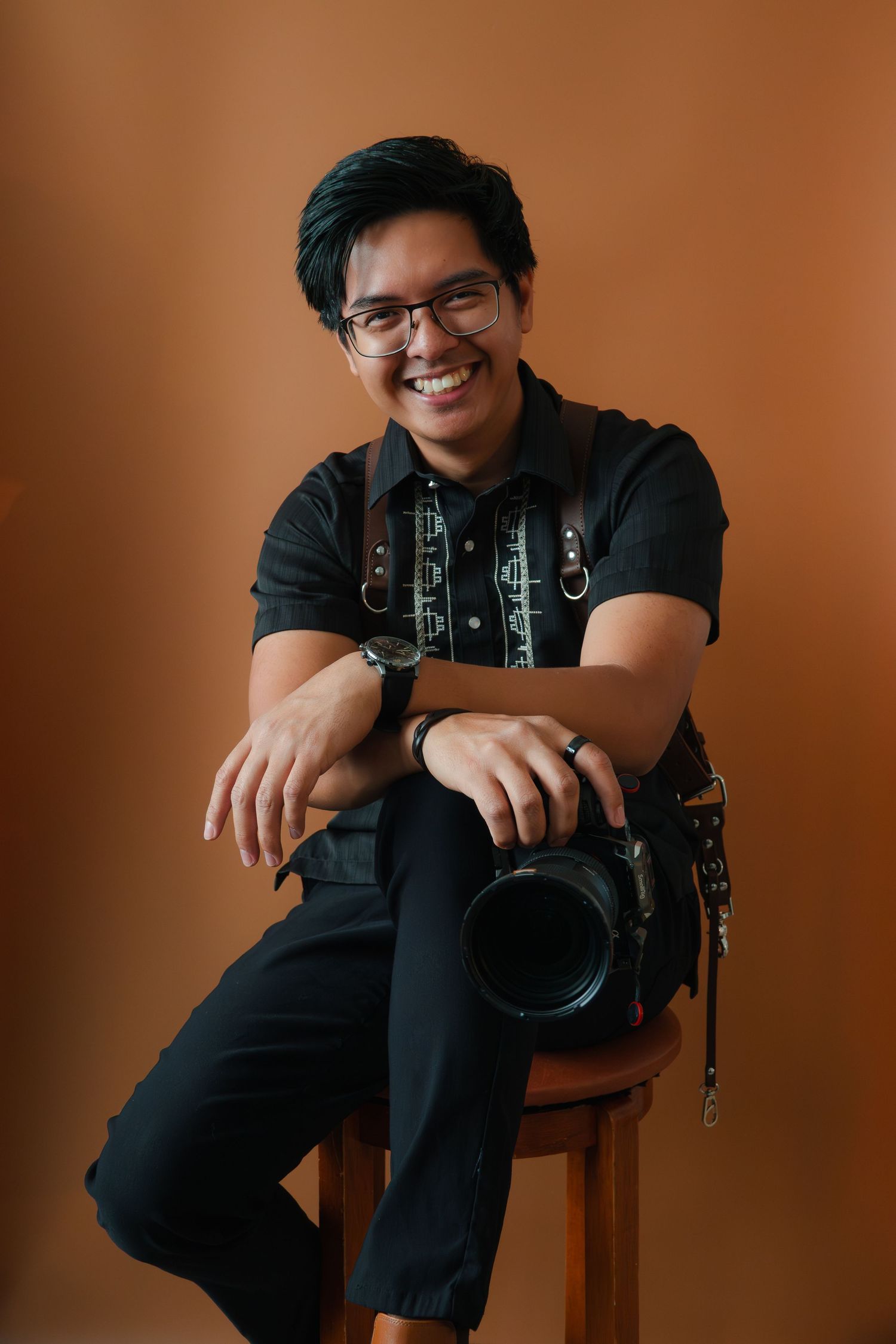 Smiling photographer sitting on stool holding DSLR camera, wearing black outfit and glasses against brown background.