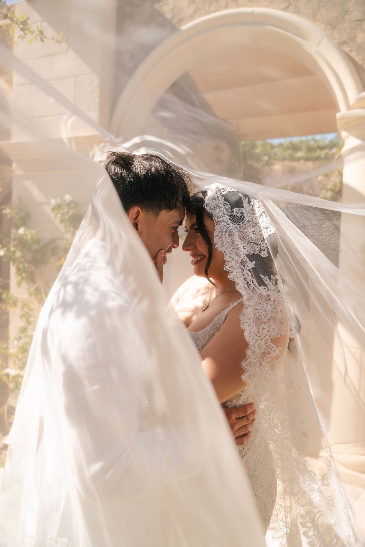 Bride and groom share an intimate moment wrapped together under a flowing lace wedding veil.