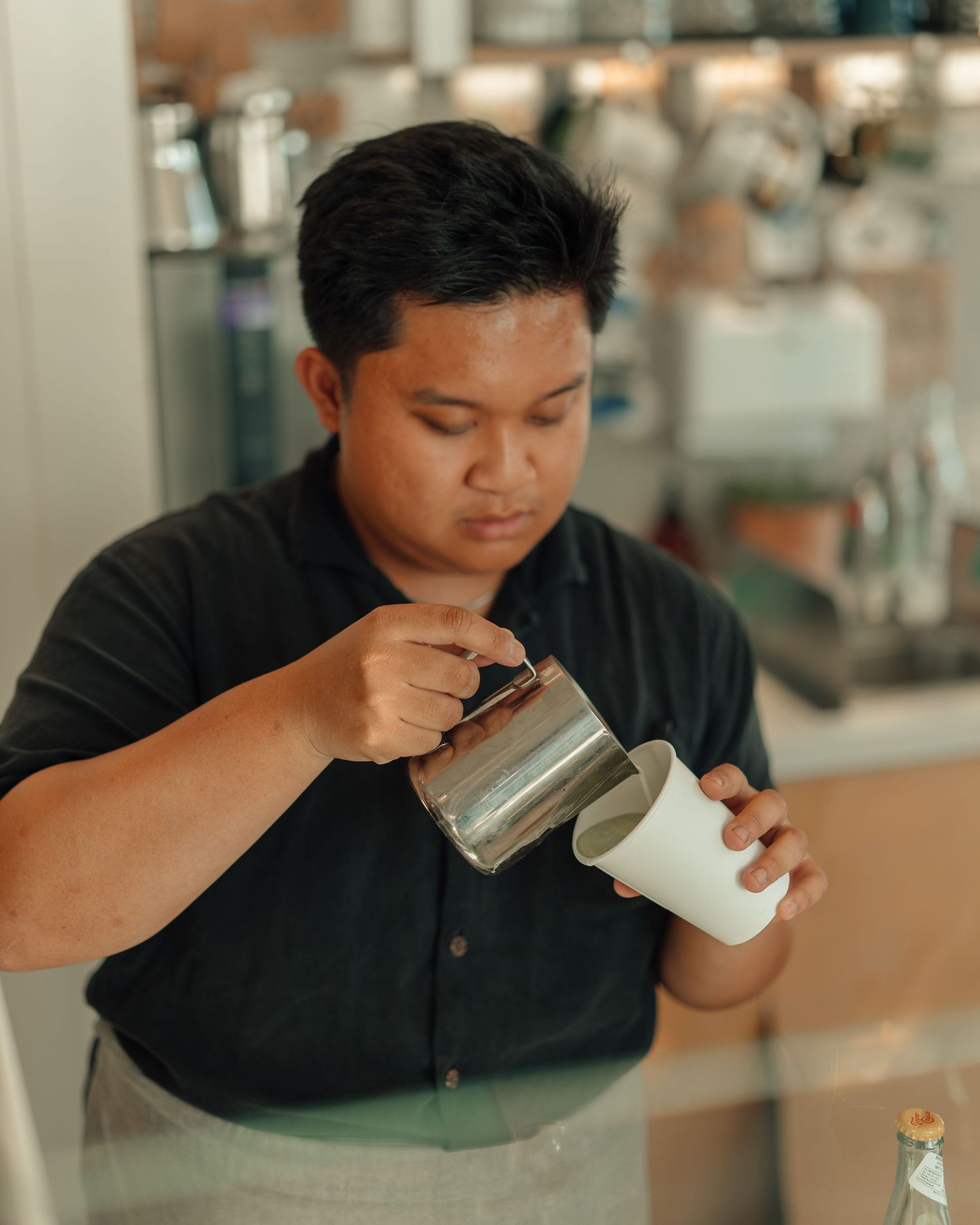 Barista pouring steamed milk from pitcher into white cup at specialty coffee bar.