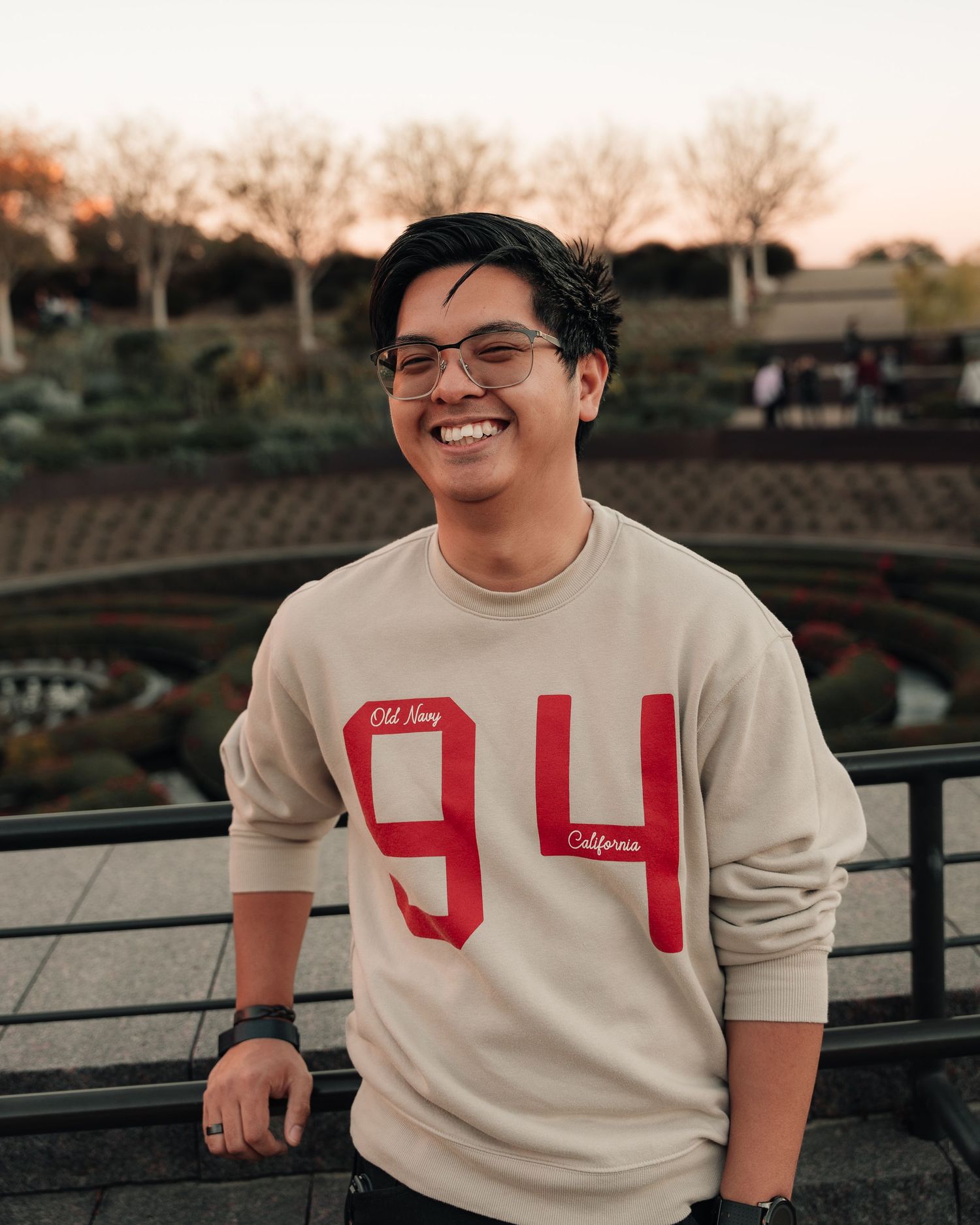 Smiling young man wearing a beige 94 California sweatshirt, standing outdoors with trees and a bridge in the background.