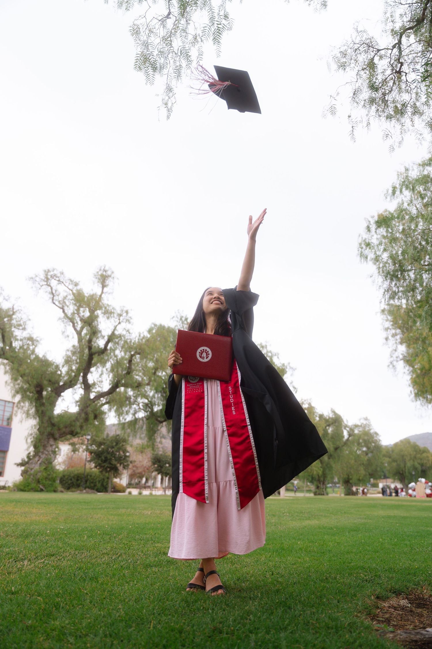 Graduate in black gown with red stole tossing mortarboard cap on green campus lawn.