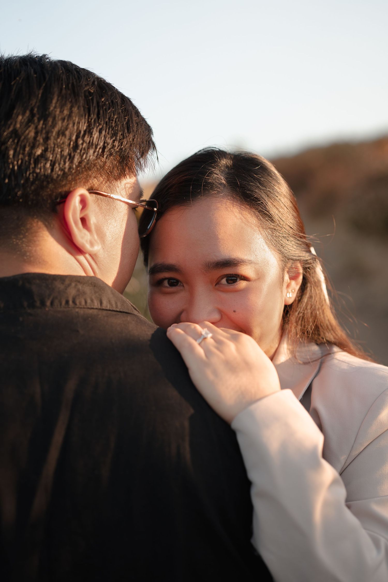 Woman smiling shyly behind her hand adorned with an engagement ring while embracing her partner outdoors.