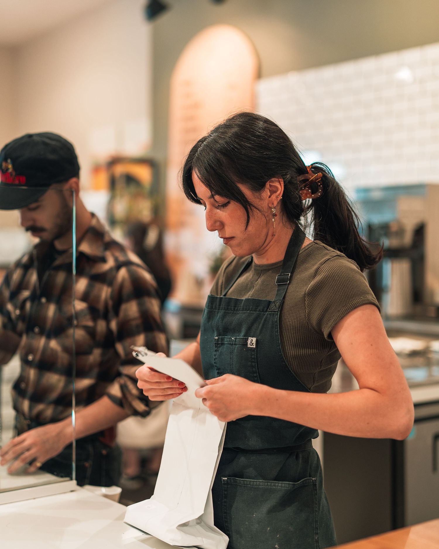 Female barista in green apron packing a takeout order at a coffee shop counter.