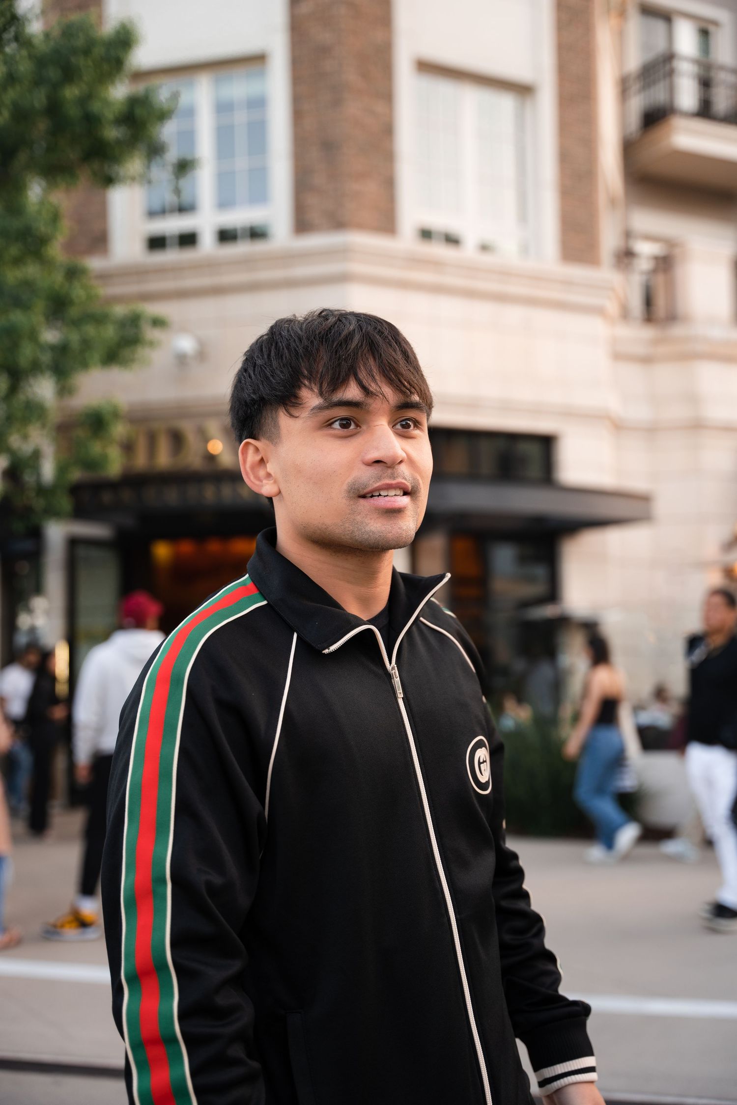 Young man in black Gucci tracksuit smiling on a busy urban street.